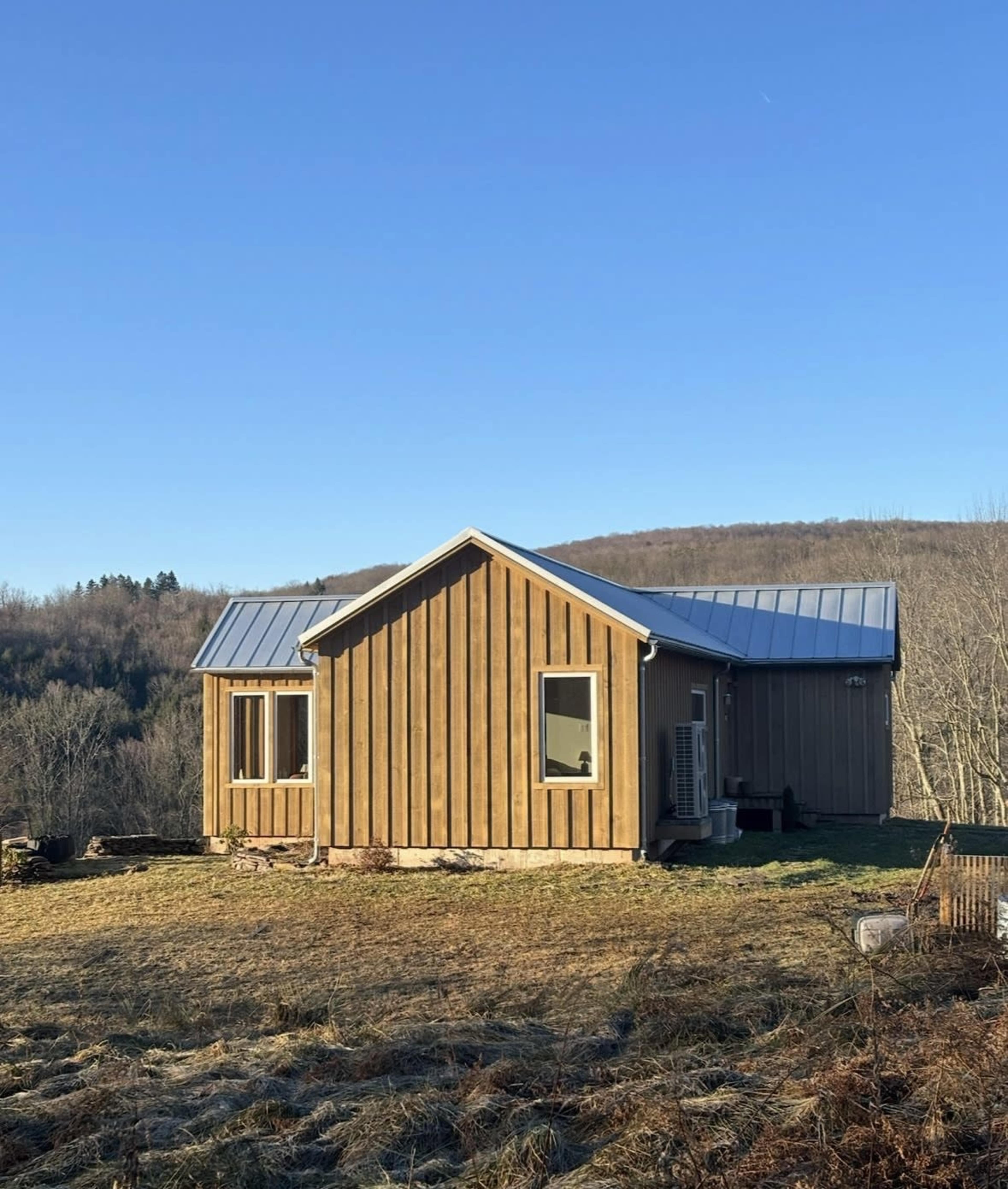 A wooden cabin with a metal roof is set on a grassy hillside with trees in the background.