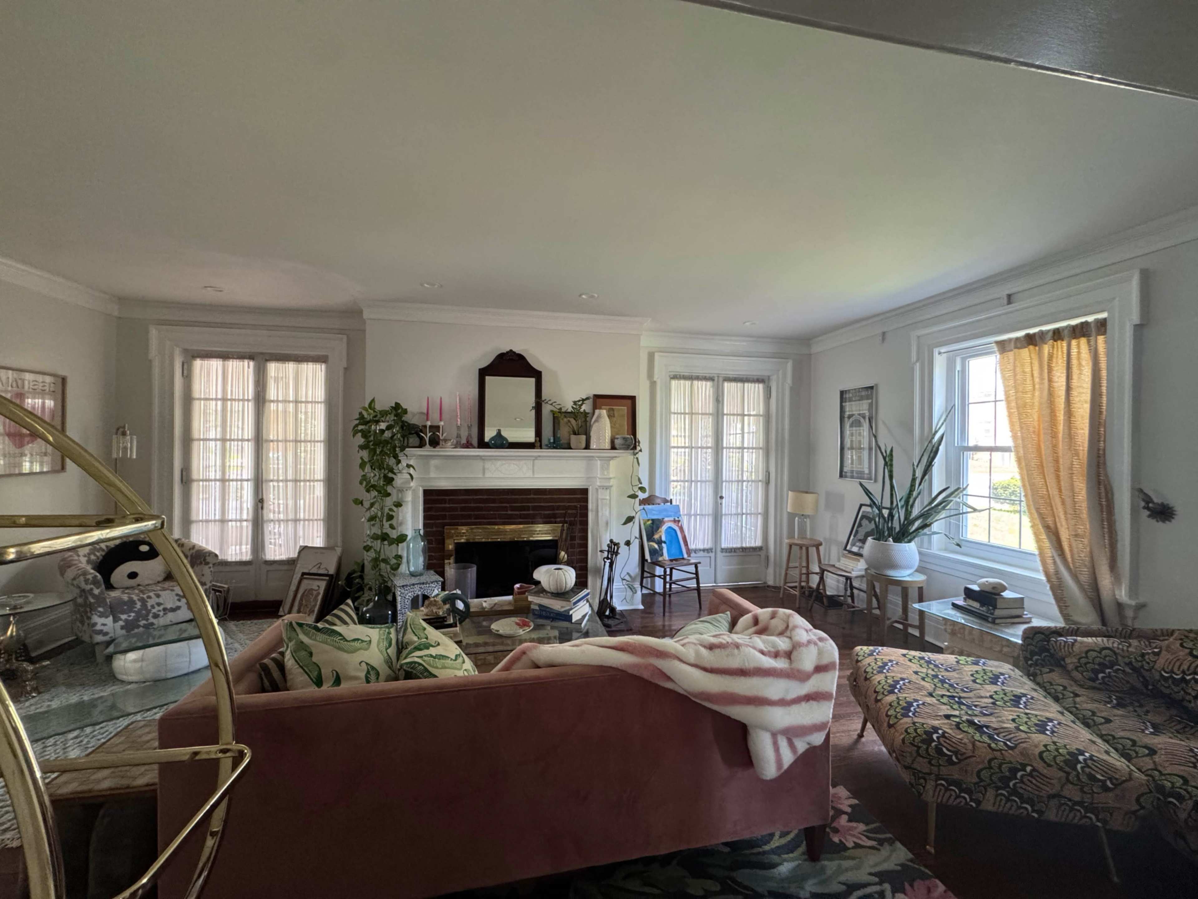 The living room features a pink sofa, a fireplace, various plants, and a mix of furniture arranged around a central area.