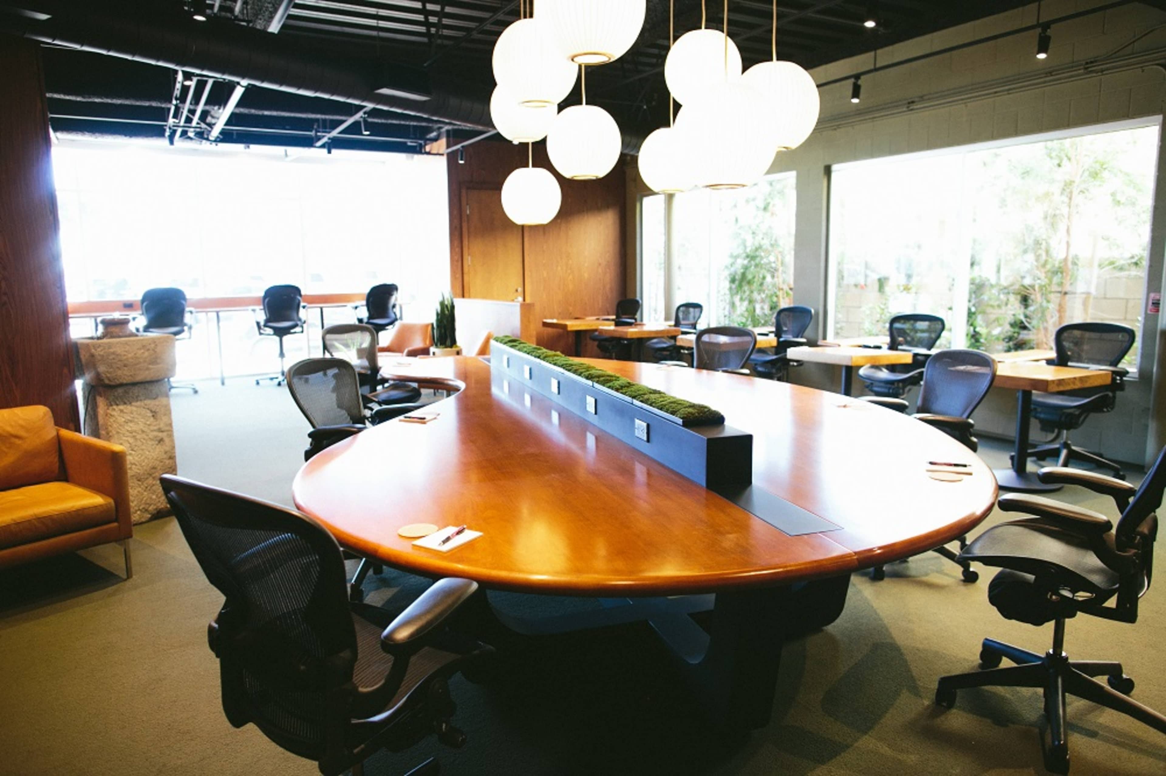 A large conference room features a round wooden table surrounded by ergonomic chairs and illuminated by modern pendant lights.