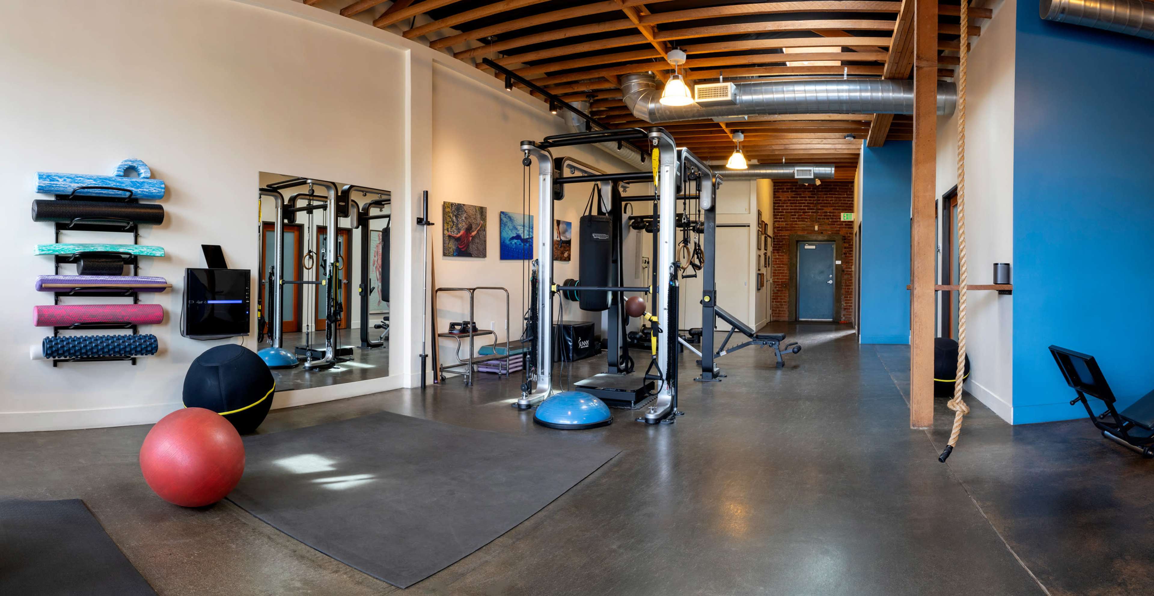 A spacious gym interior featuring various exercise equipment, including weights, stability balls, and yoga mats arranged on the walls.