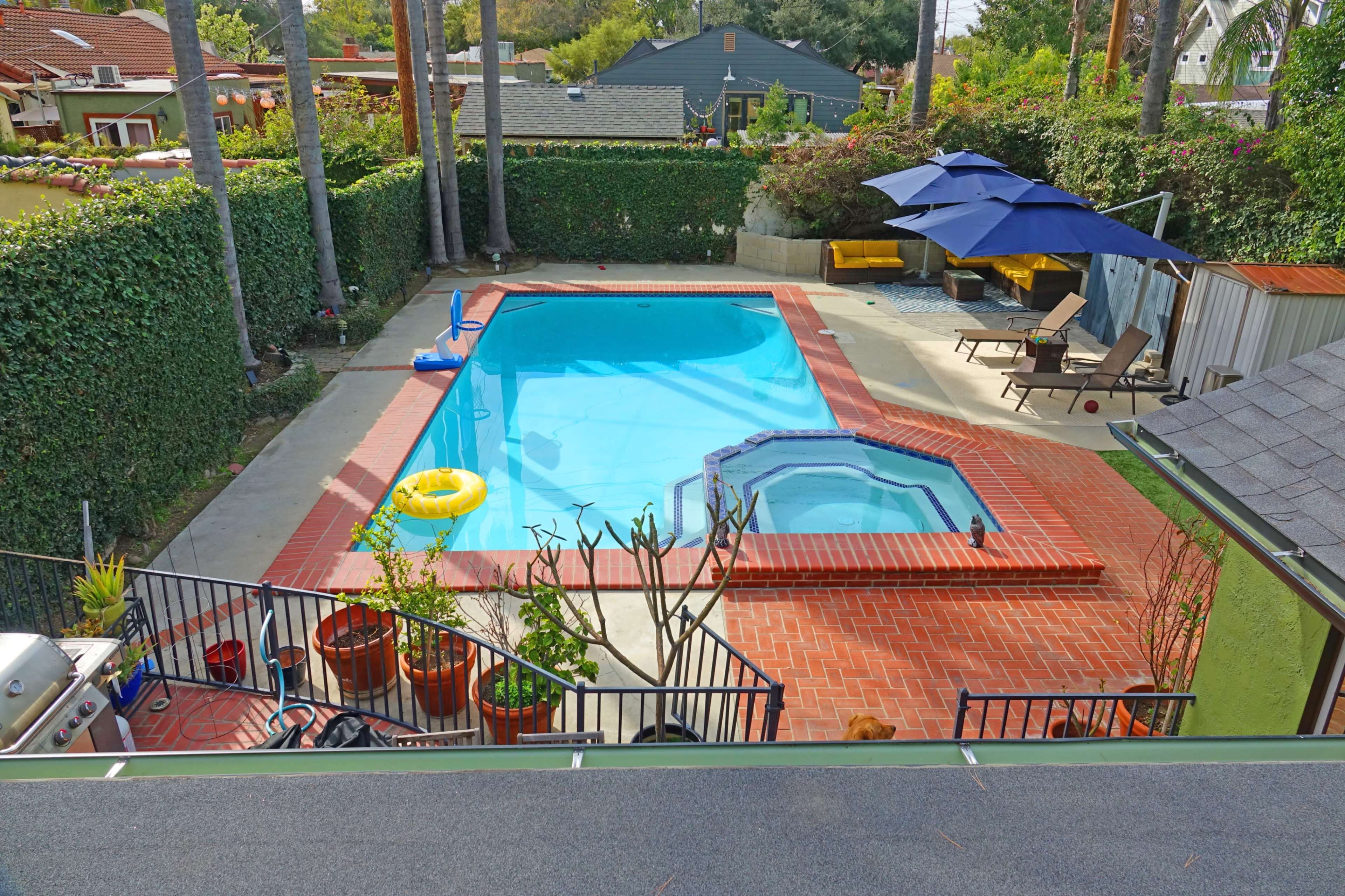 The image shows a residential backyard with a swimming pool, a hot tub, lounge chairs, an umbrella, and potted plants surrounded by a hedge.