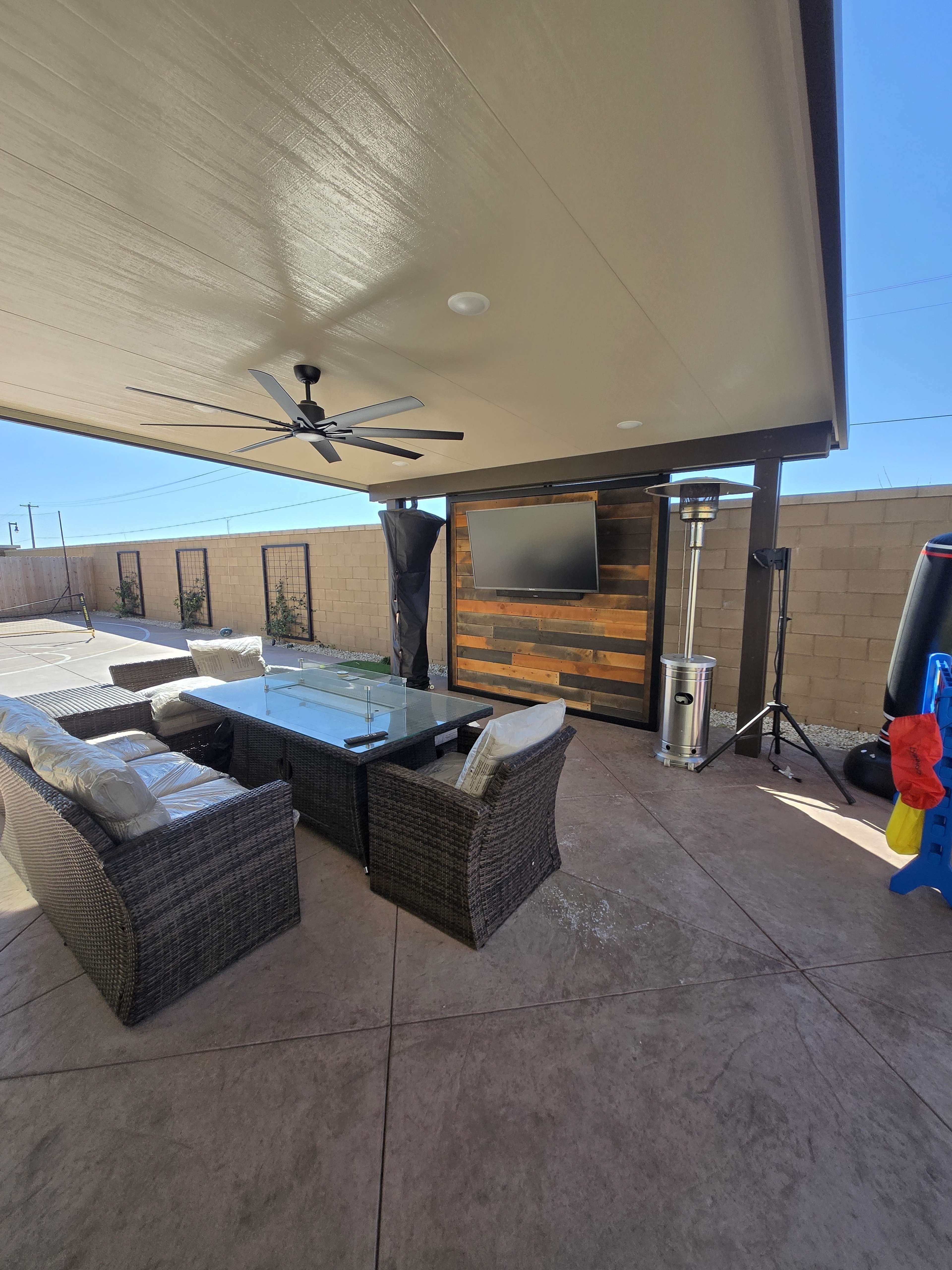 The image shows a covered outdoor seating area with a sofa set, a coffee table, an outdoor heater, and a wall-mounted television.