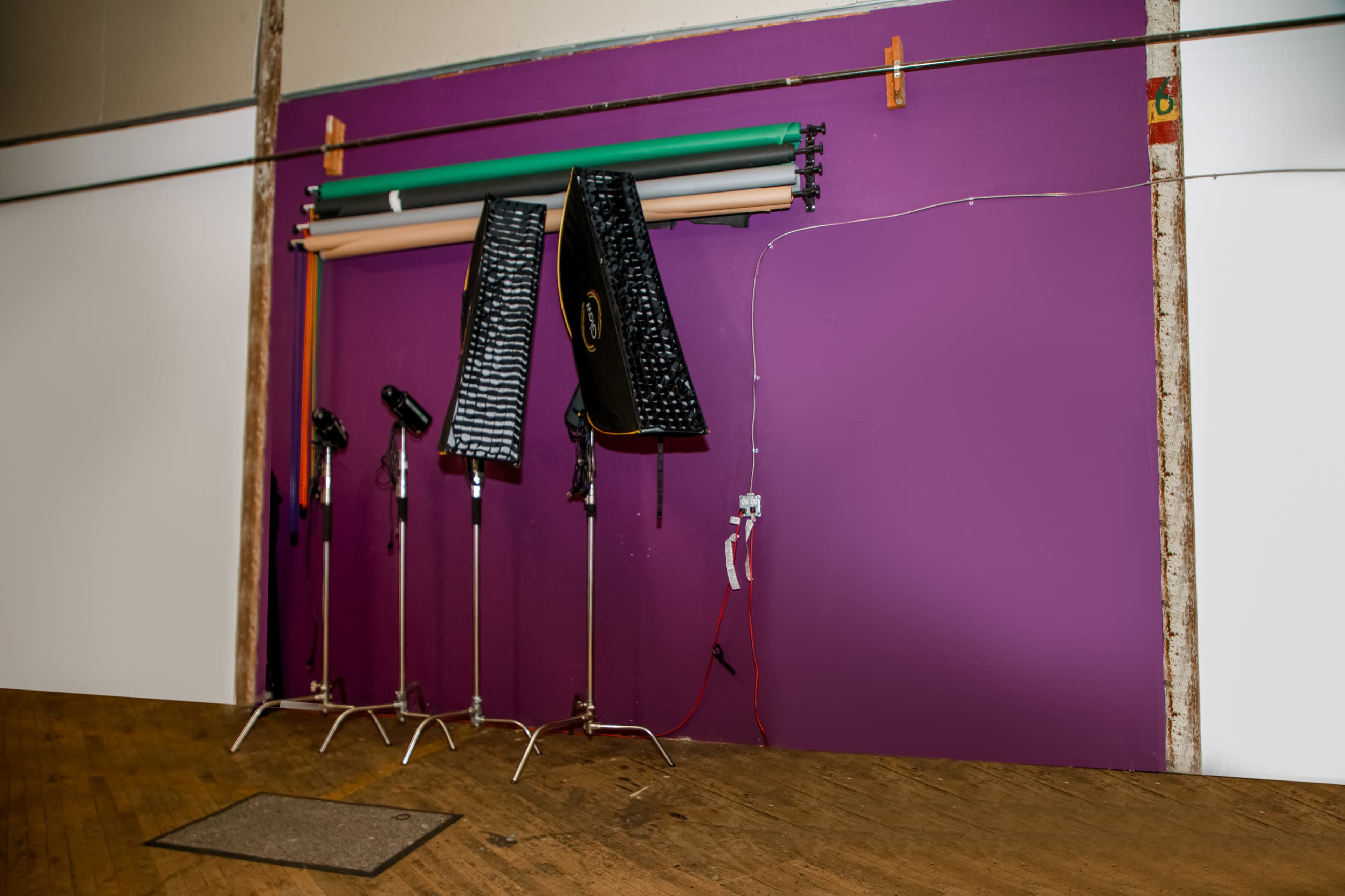 The image shows a purple wall with several photography lights and backdrop rolls mounted above a wooden floor.