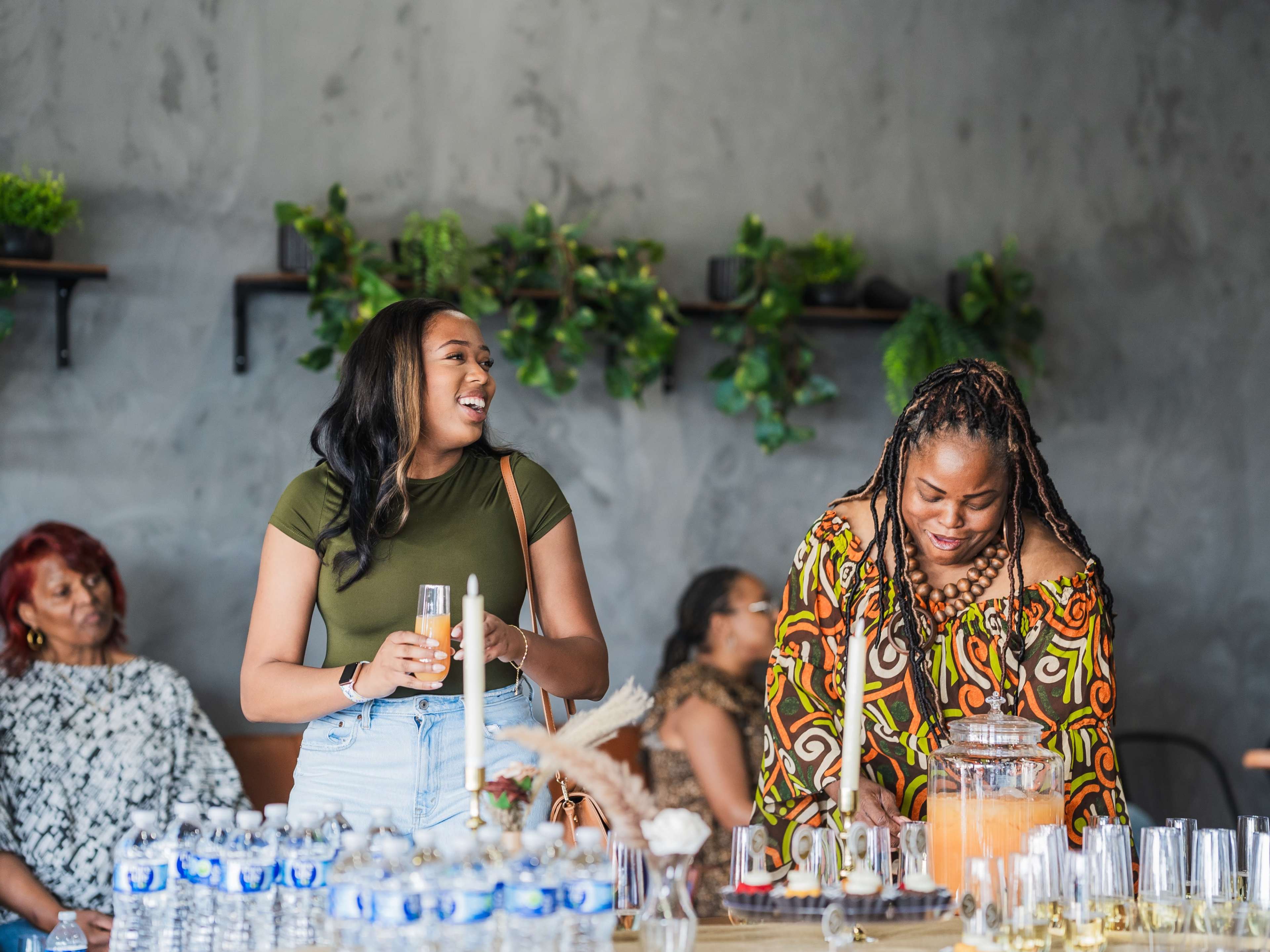 A group of women gathers in a modern interior space with greenery on the walls, while two women engage in conversation near a table with drinks and snacks.