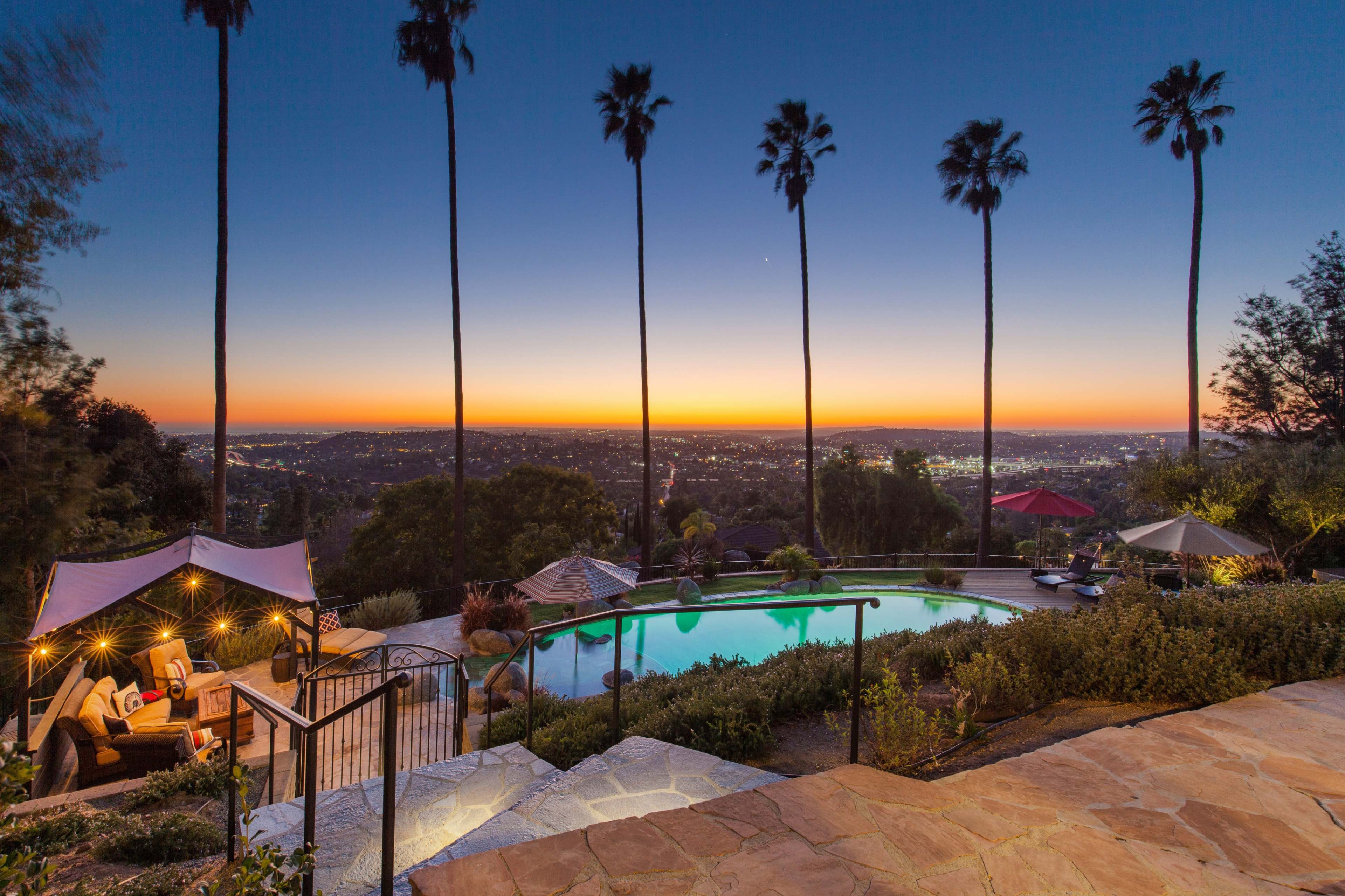 A swimming pool surrounded by palm trees overlooks a city skyline during sunset.