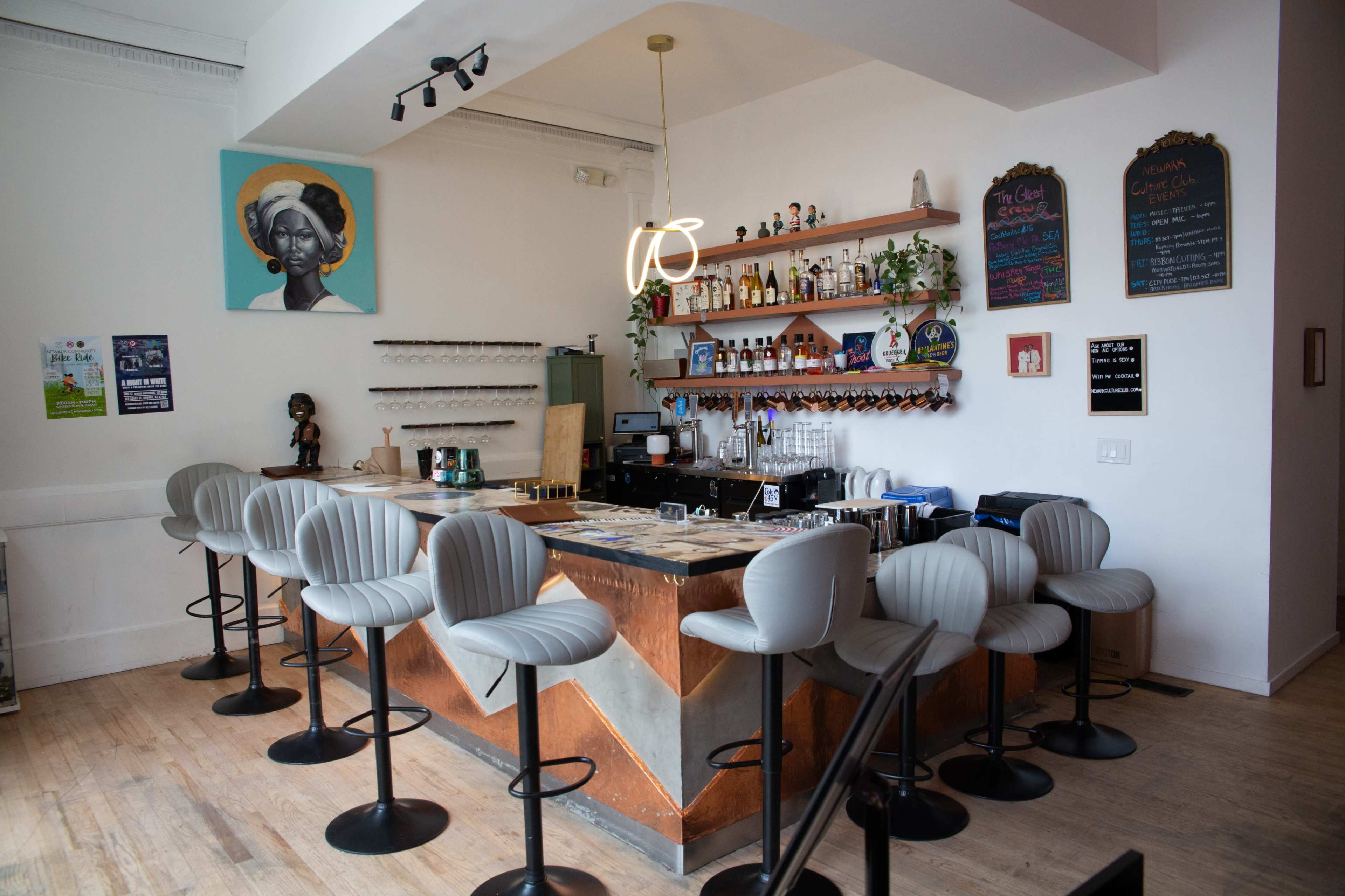 The image shows a modern bar area with a curved counter, high stools, and a wall adorned with bottles and artwork.