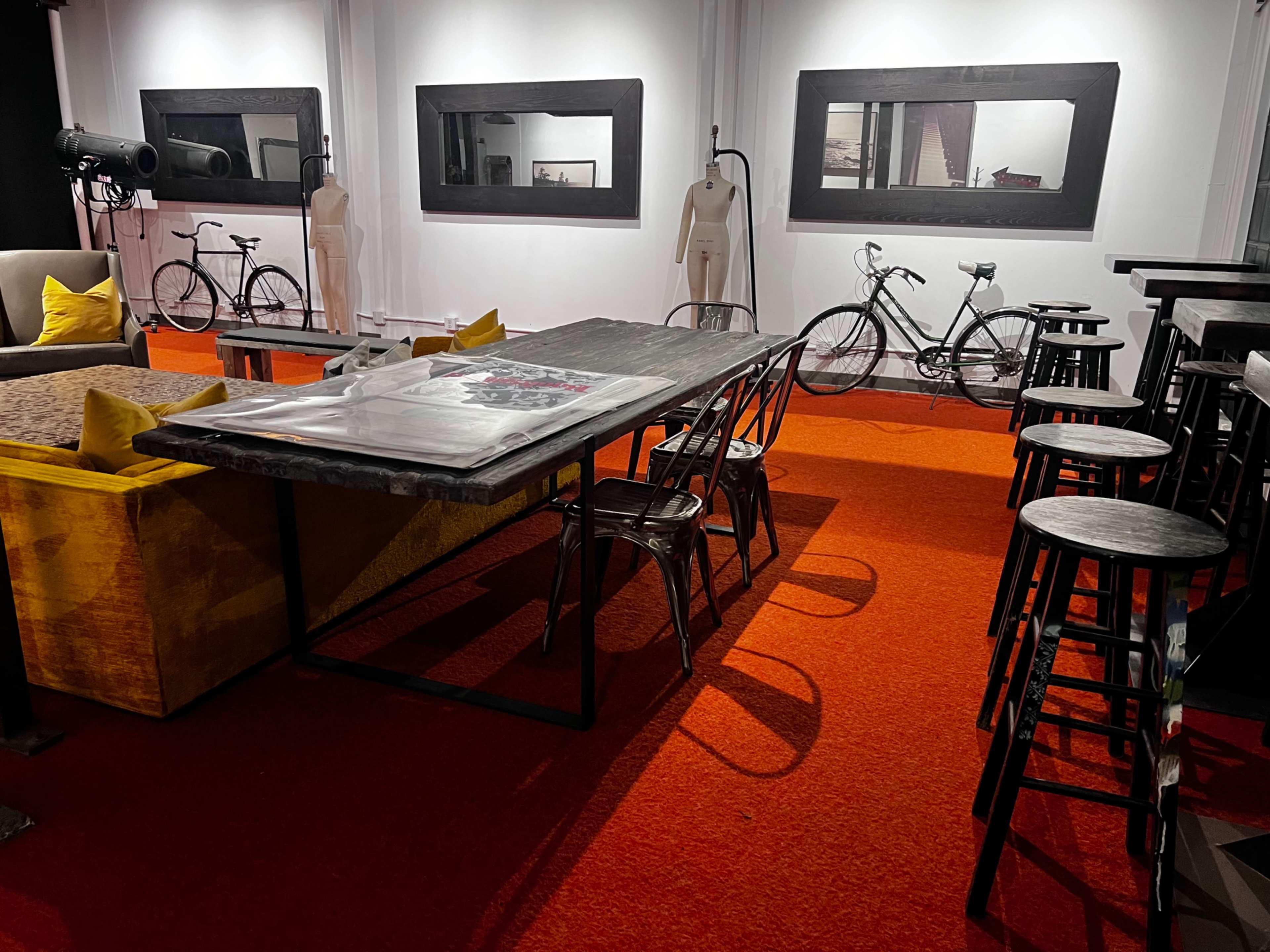 The scene features an interior space with a large wooden table surrounded by metal stools, set against an orange carpet, while vintage bicycles and framed artworks adorn the walls.