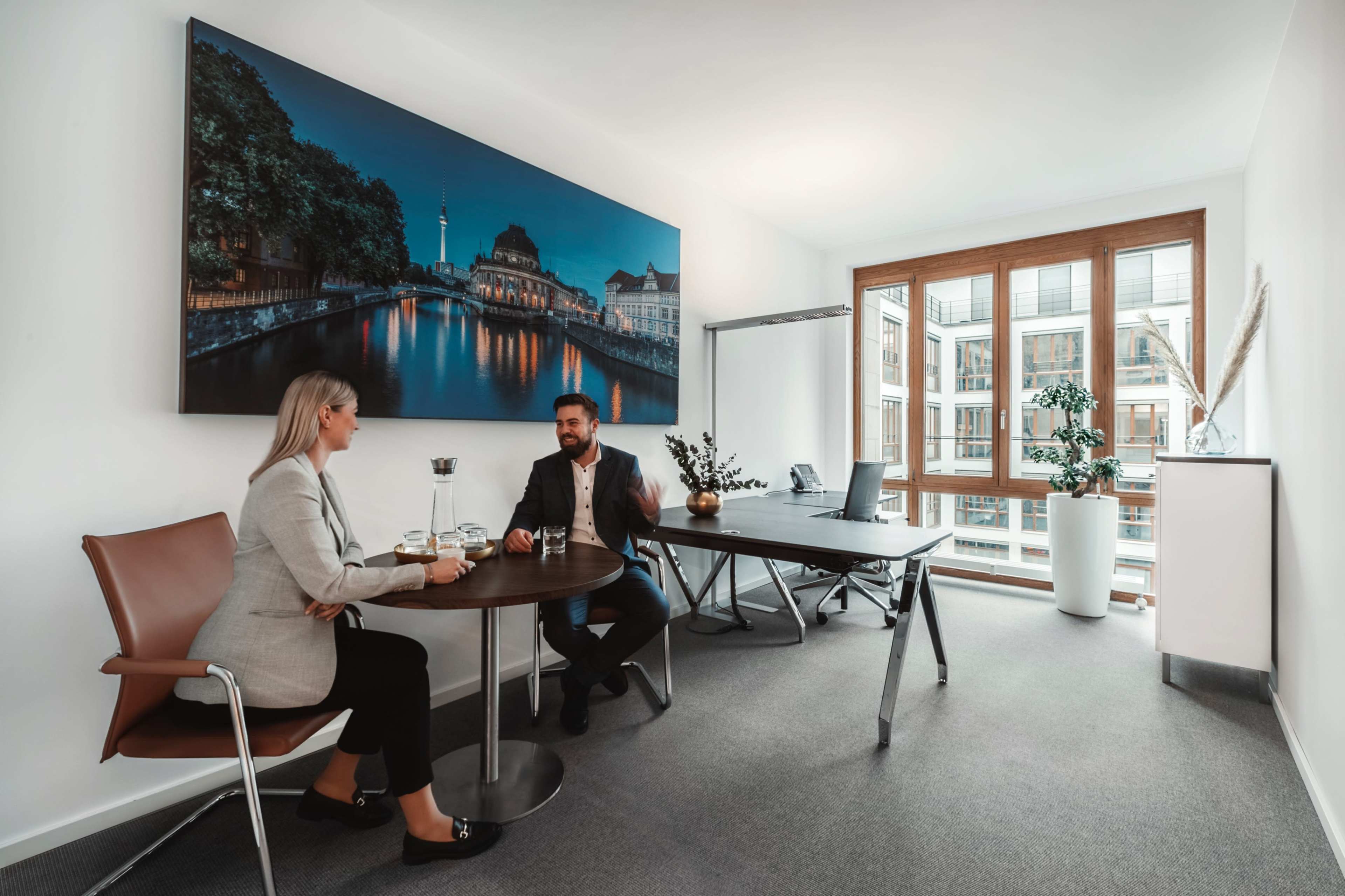 Two professionals sit at a round table in an office, conversing over drinks while a large cityscape photograph is displayed on the wall.