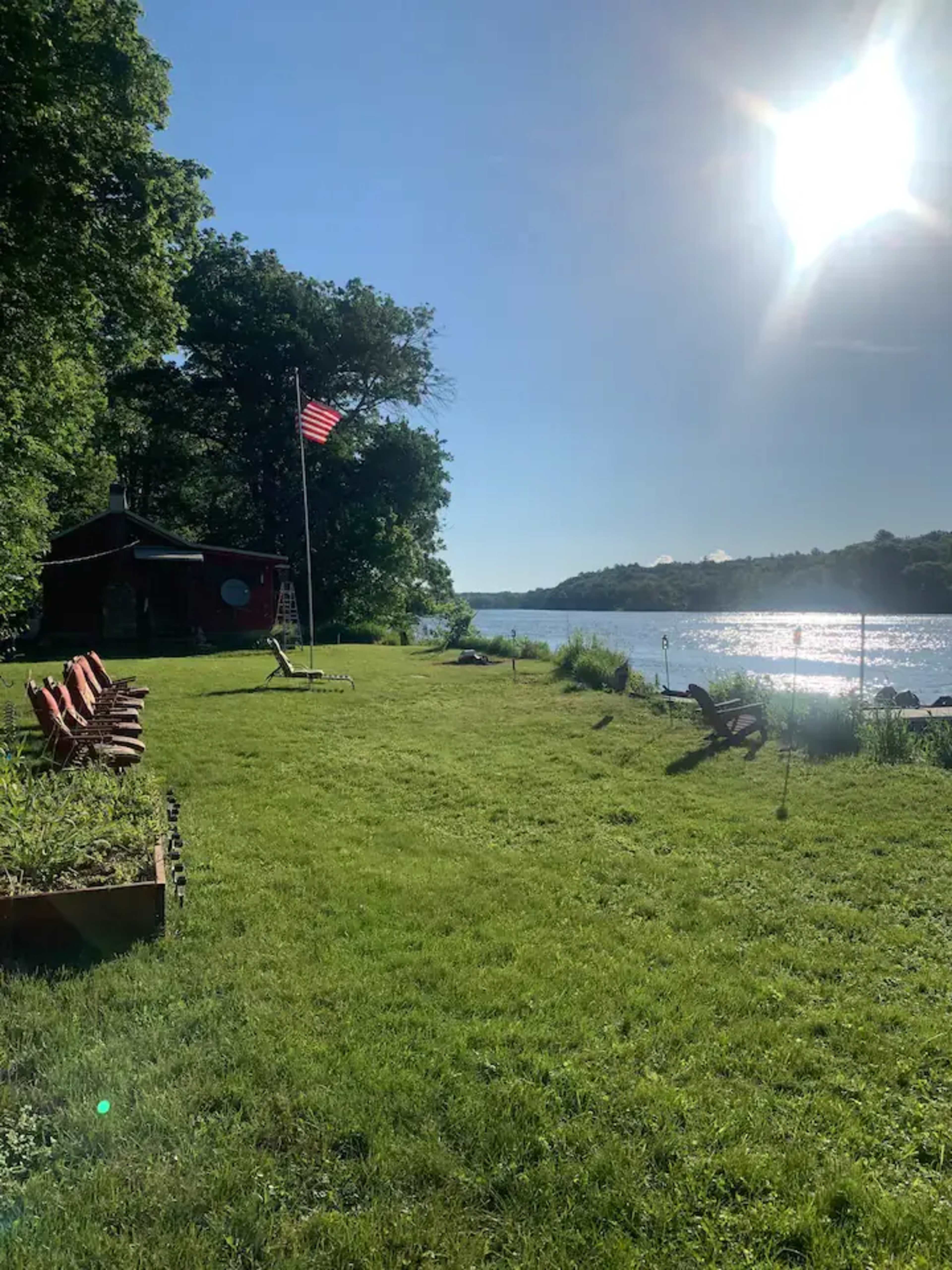 A red cottage with an American flag stands by a river, surrounded by a grassy area and chairs.