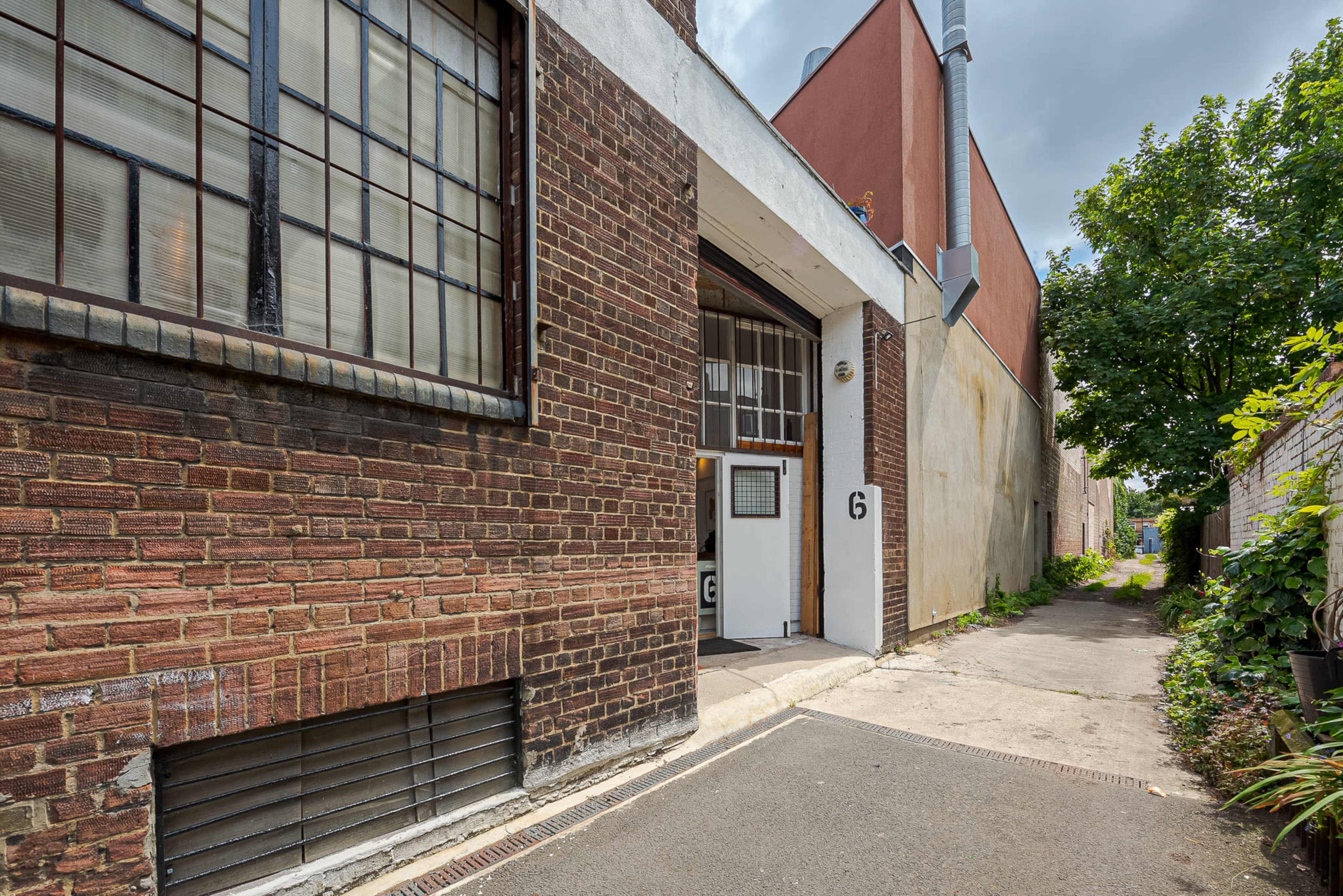 A narrow alleyway beside a brick building with large windows leading to a white entrance door marked with the number 6.