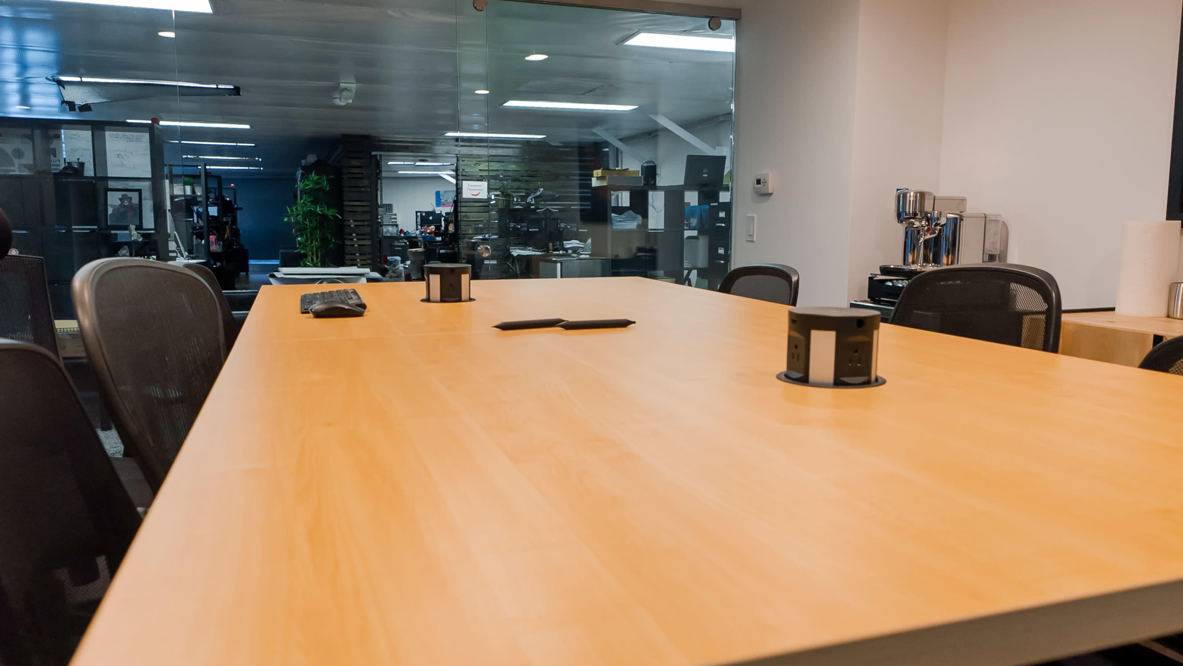 A large wooden conference table is positioned in a modern office space, with a glass partition and various office equipment visible in the background.