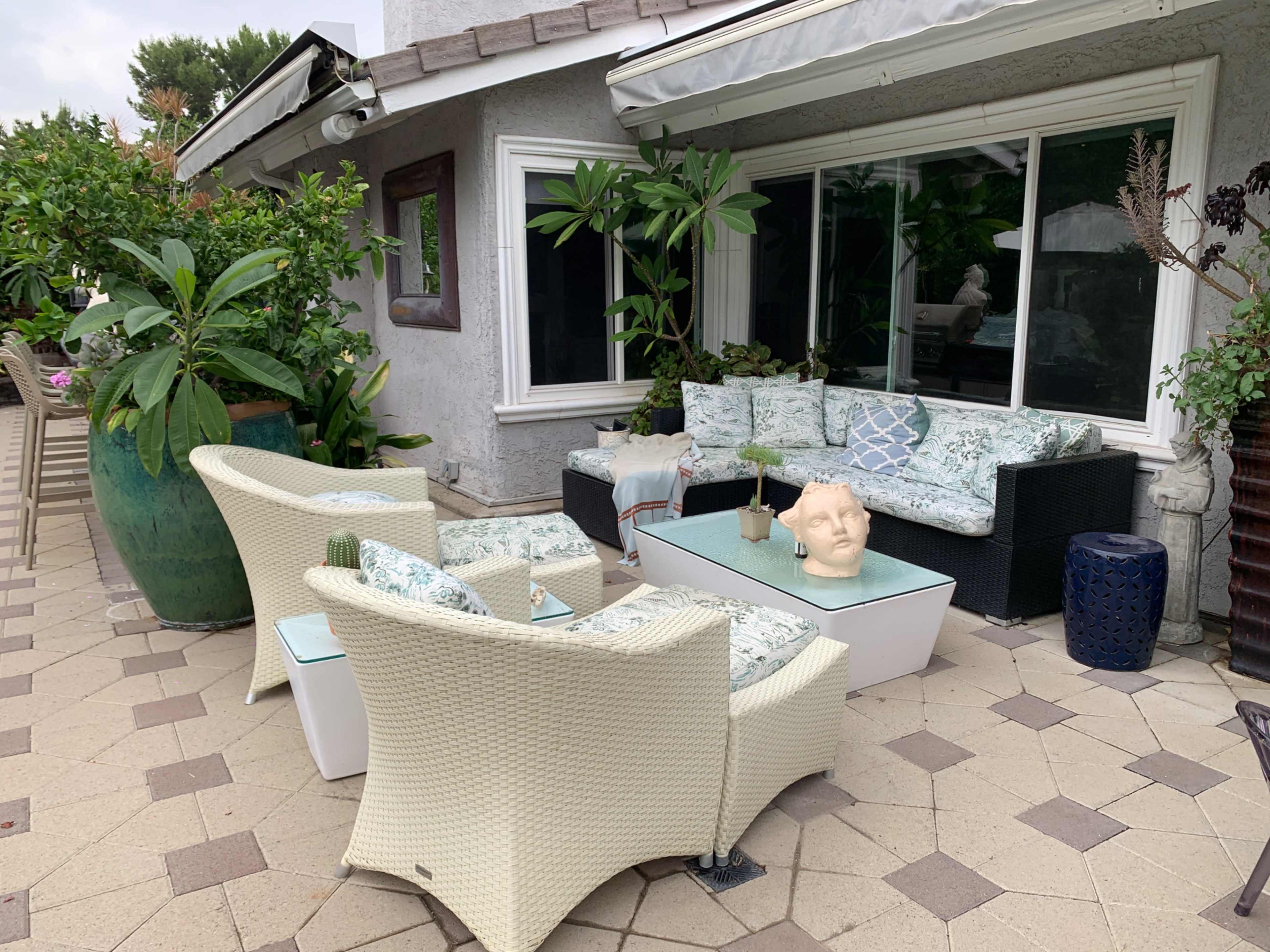 A patio setting features white wicker furniture arranged around a glass coffee table, with greenery and decorative elements in a residential outdoor space.