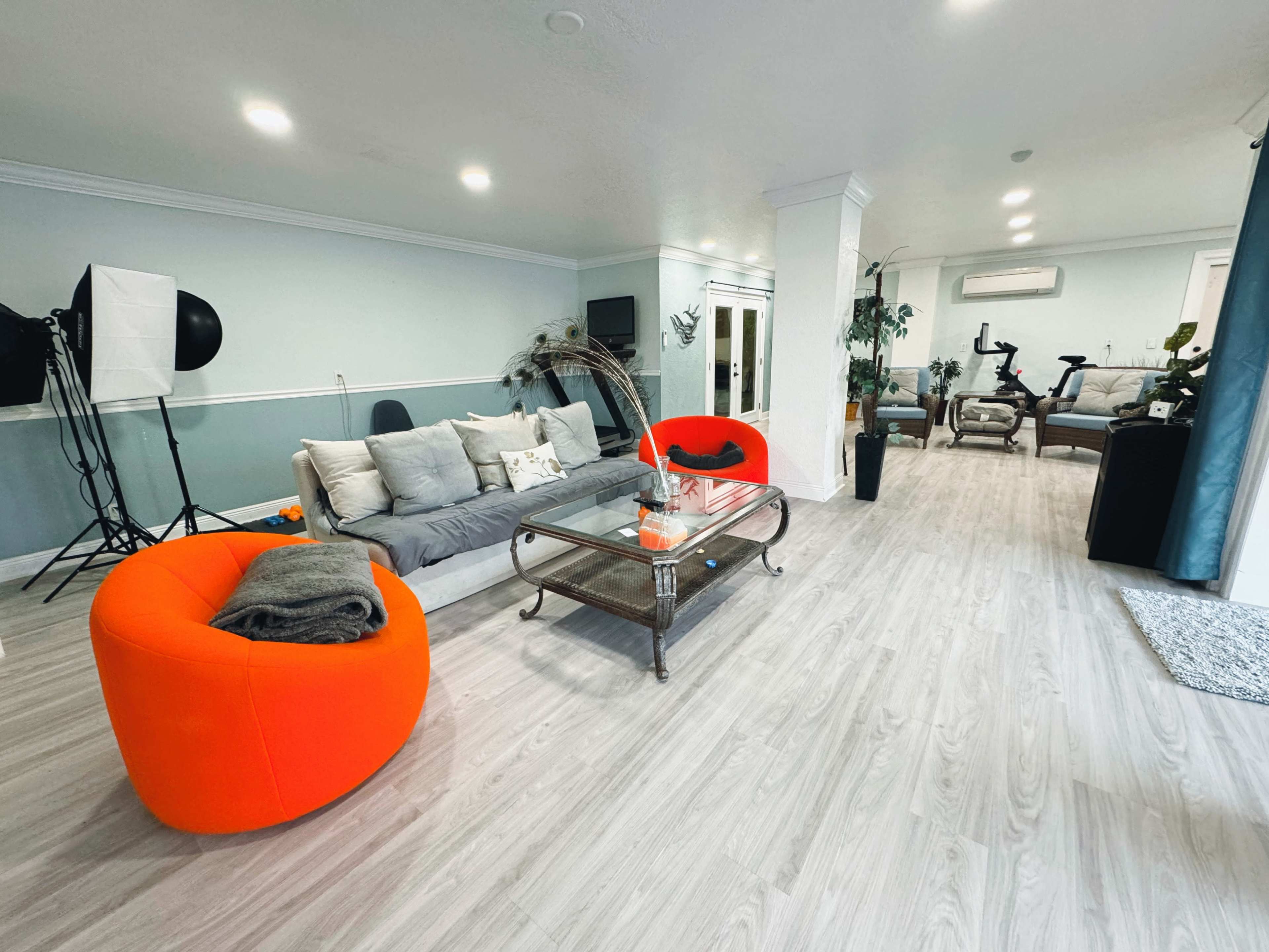 A spacious living room featuring a gray sofa, two bright orange chairs, a glass coffee table with decorative items, and exercise equipment in the background.