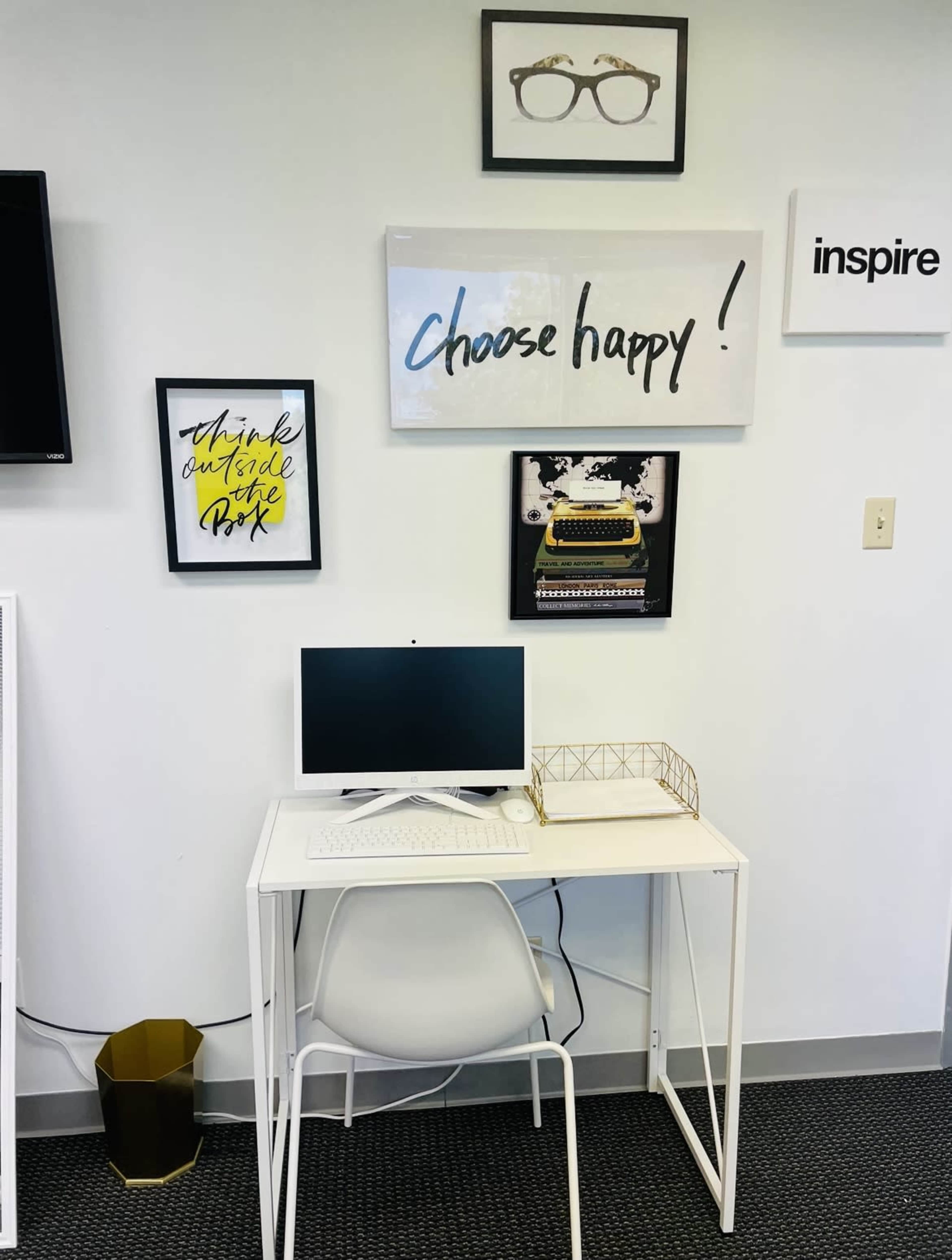 A white desk with a computer and decorative items is positioned against a wall featuring framed art and a television.