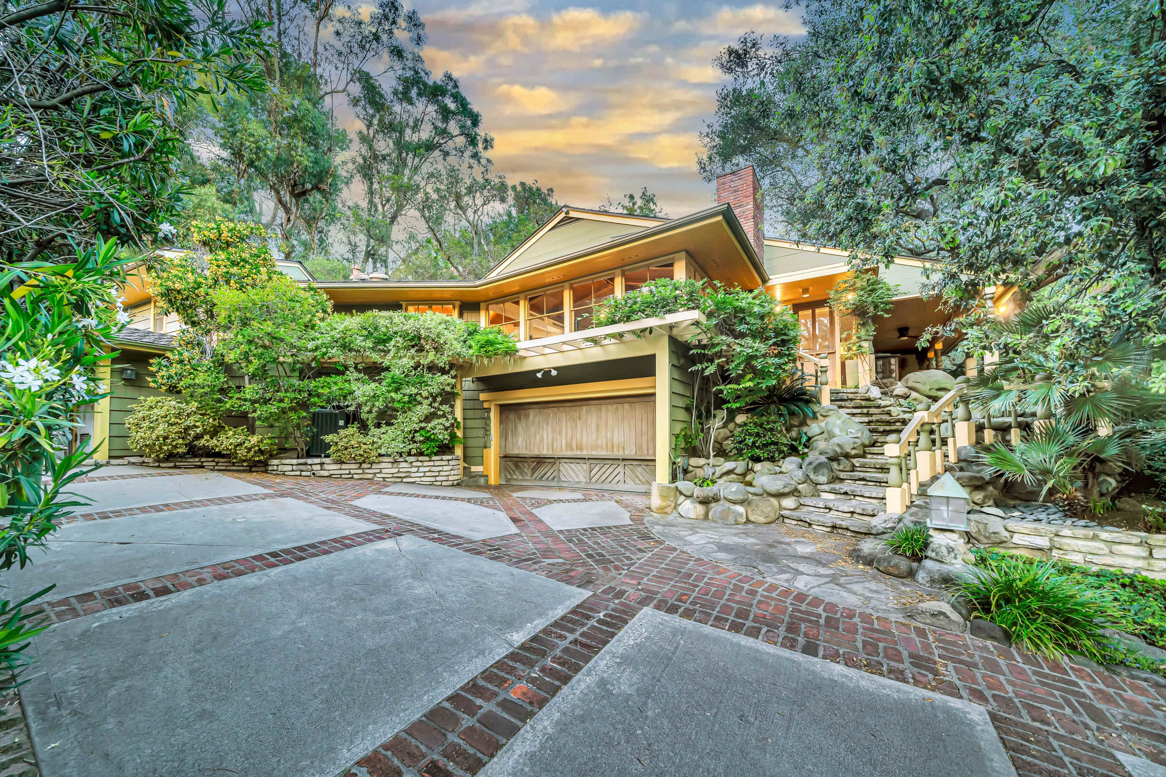 A multi-level house with a stone path and lush greenery is surrounded by trees in a tranquil setting.