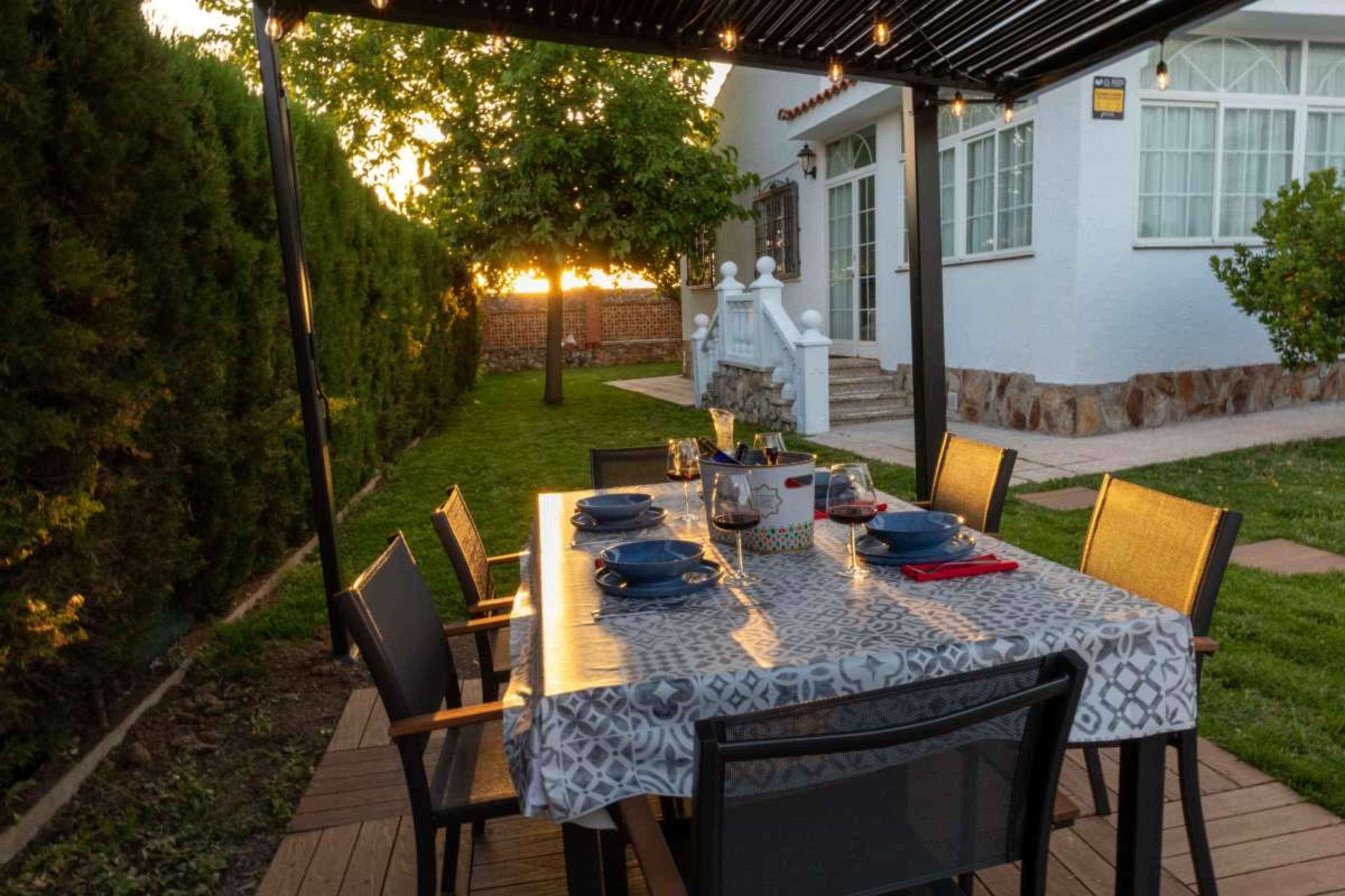 A dining table is set outdoors under a pergola, surrounded by chairs and illuminated by string lights, with a view of a garden and a house.