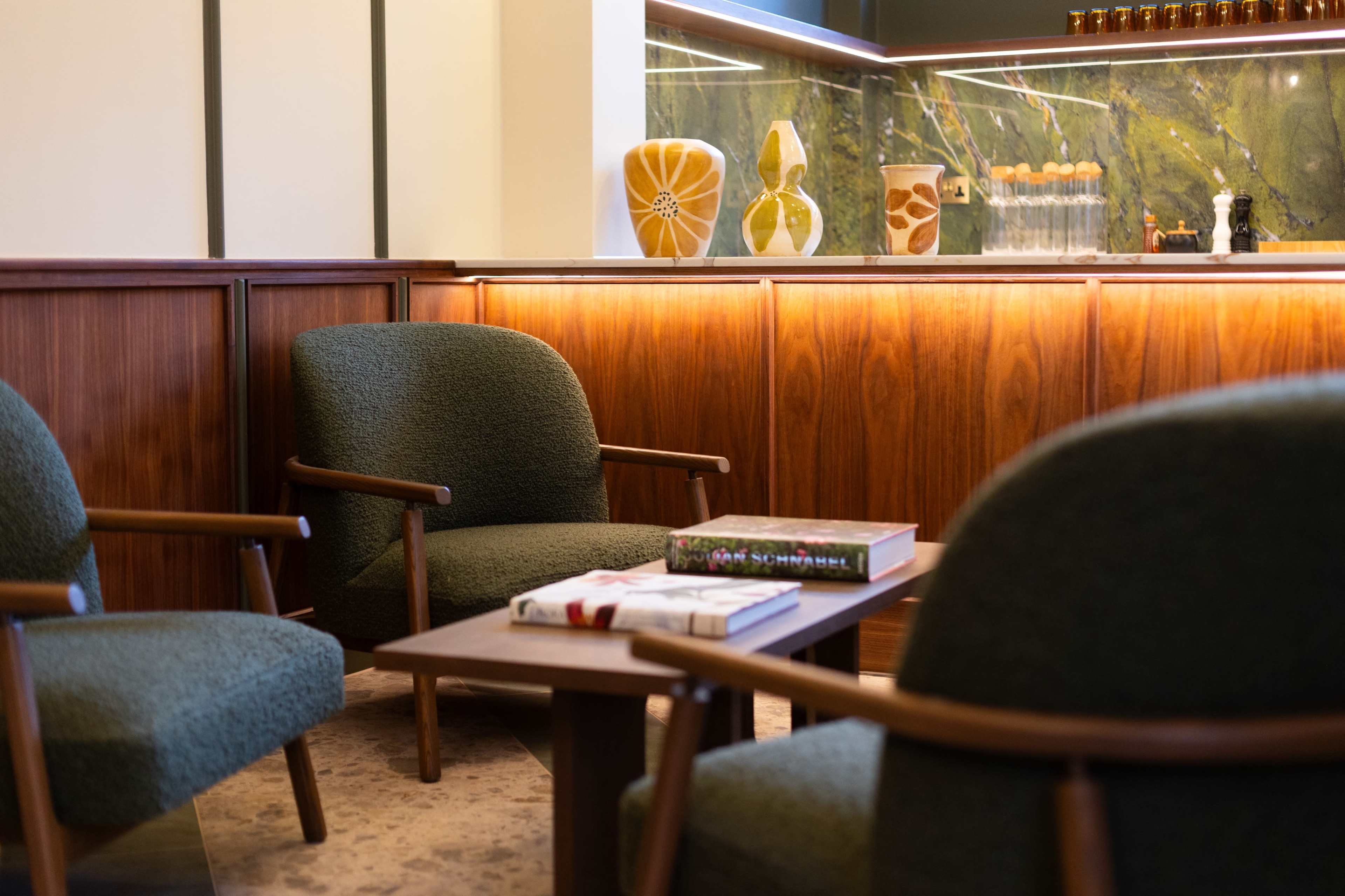 The image features a cozy seating area with three green armchairs arranged around a wooden table, set against a backdrop of a warm-lit wooden panel and decorative vases.