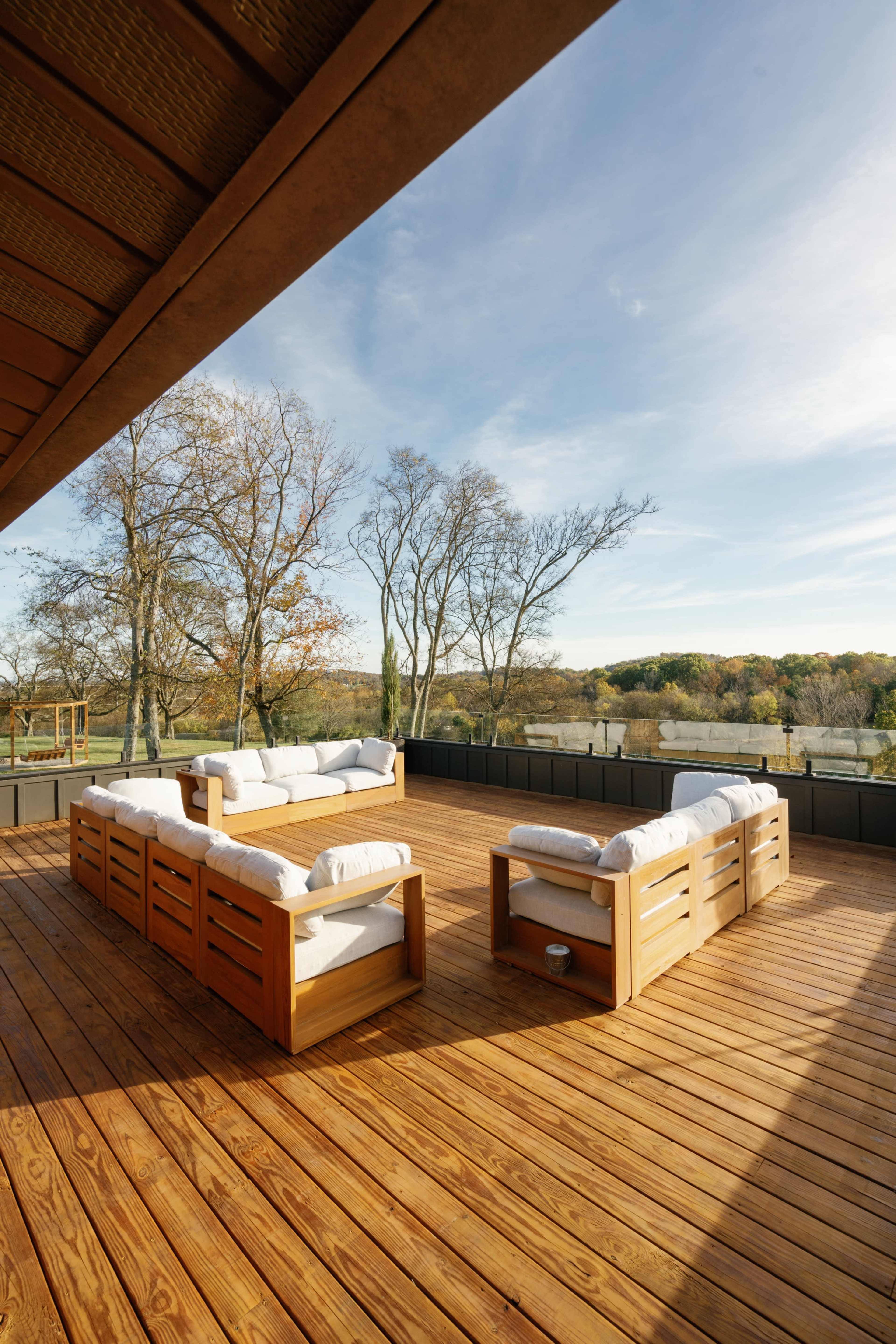 A wooden deck features two sofa sets with white cushions, overlooking a landscape of trees and hills under a clear sky.