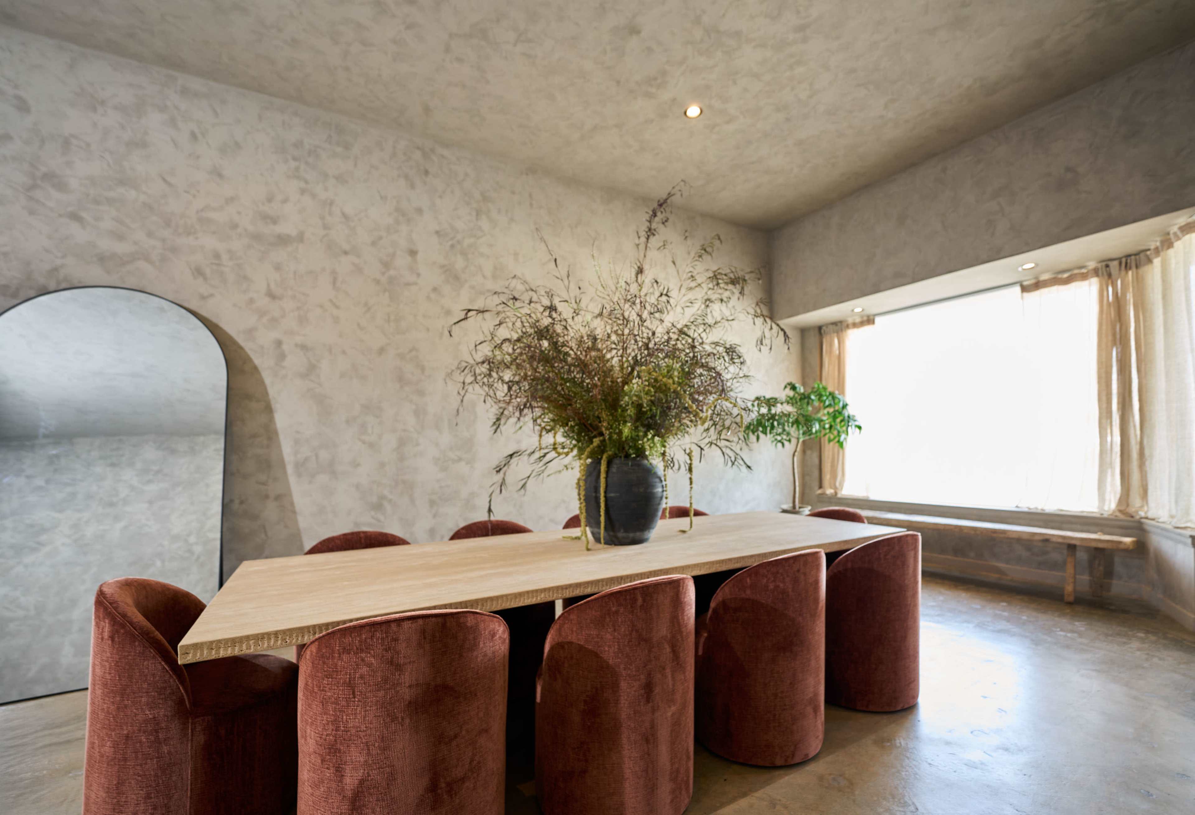 A large wooden dining table is surrounded by several upholstered chairs, with a vase of flowers as a centerpiece and a large window letting in natural light.