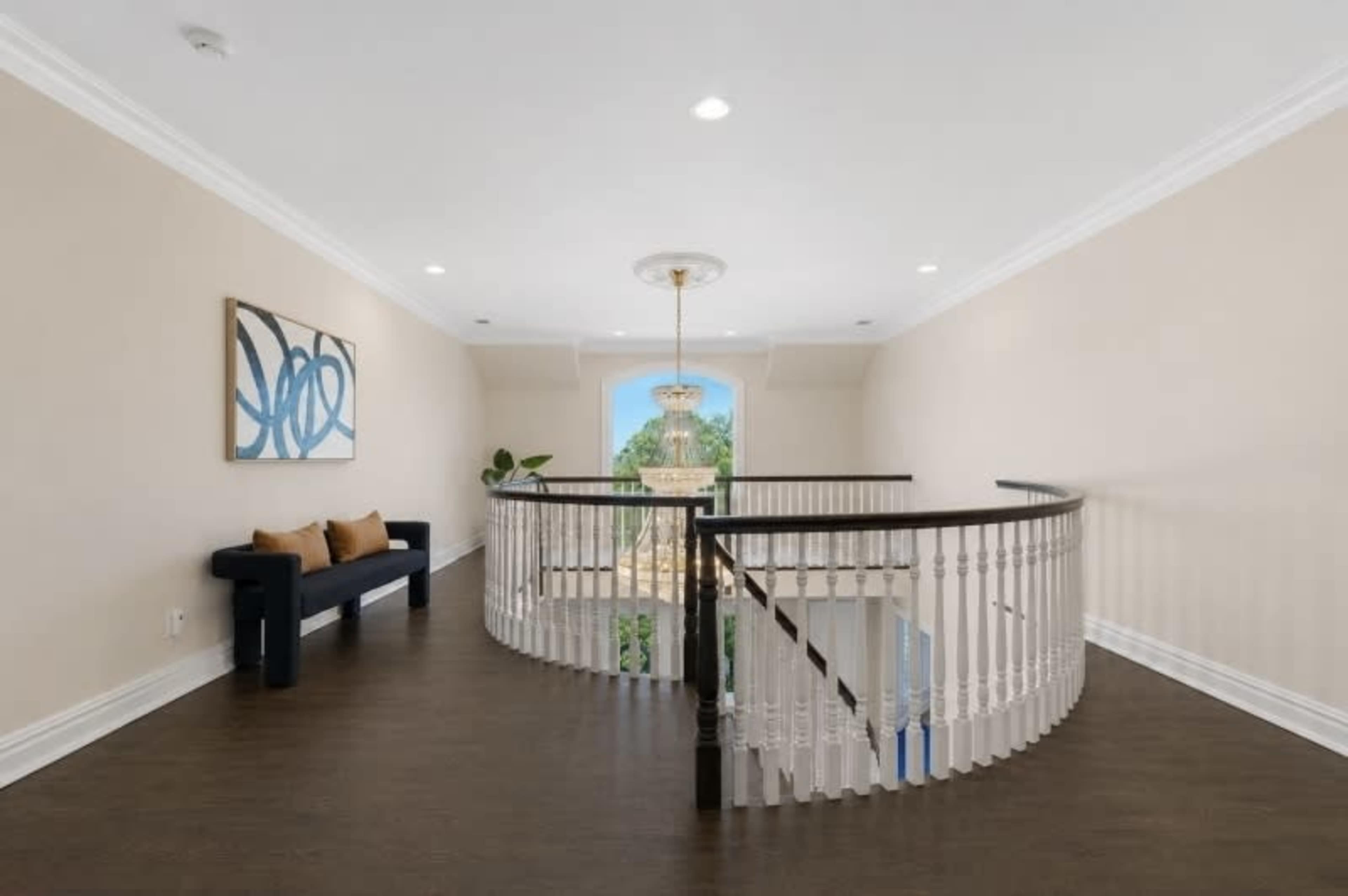 The image shows a well-lit upstairs landing with a curved staircase, a decorative chandelier, and a small seating area against a cream-colored wall.