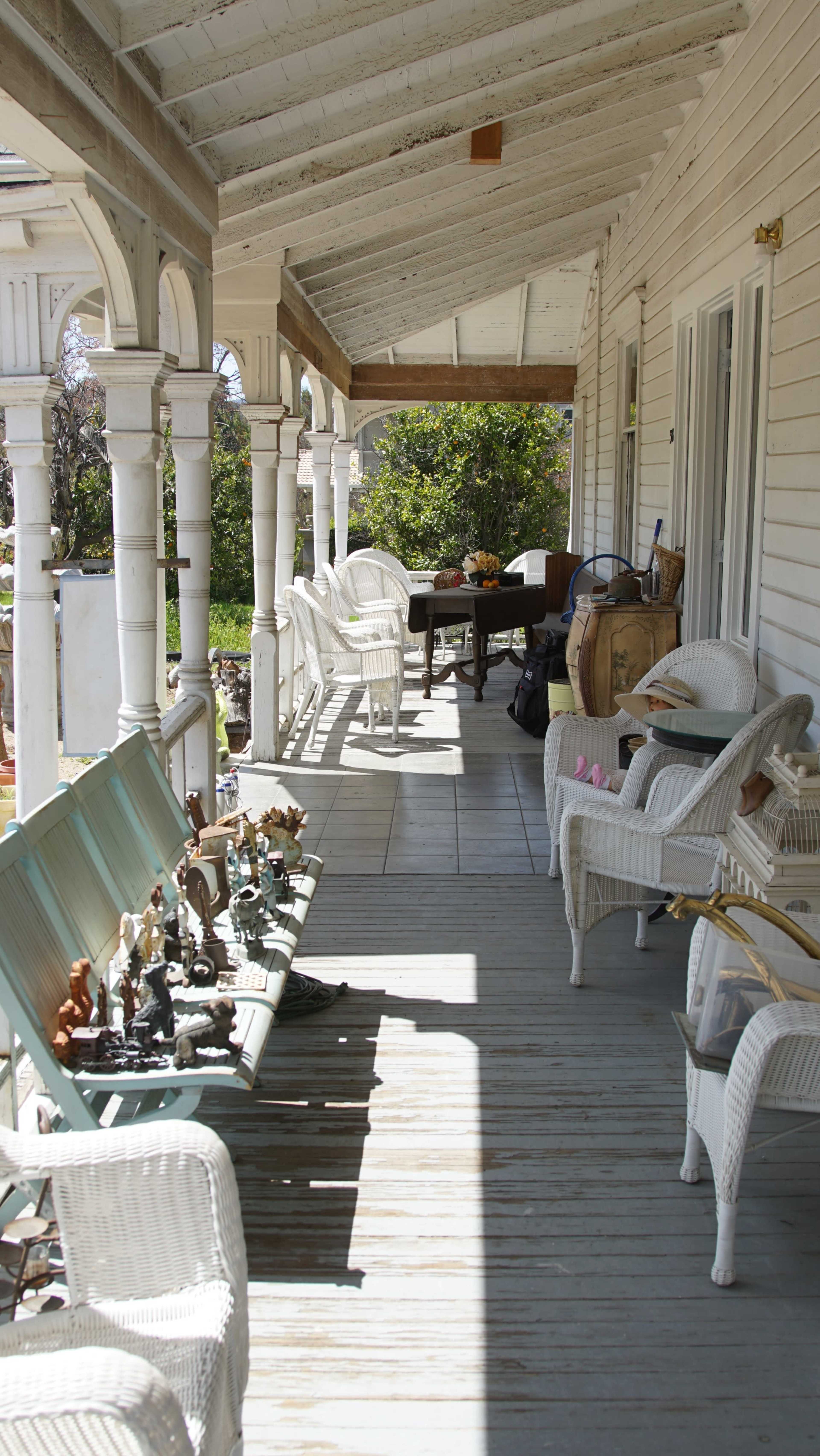 A long, covered porch features several white wicker chairs and a table, with decorative figurines arranged along one side.