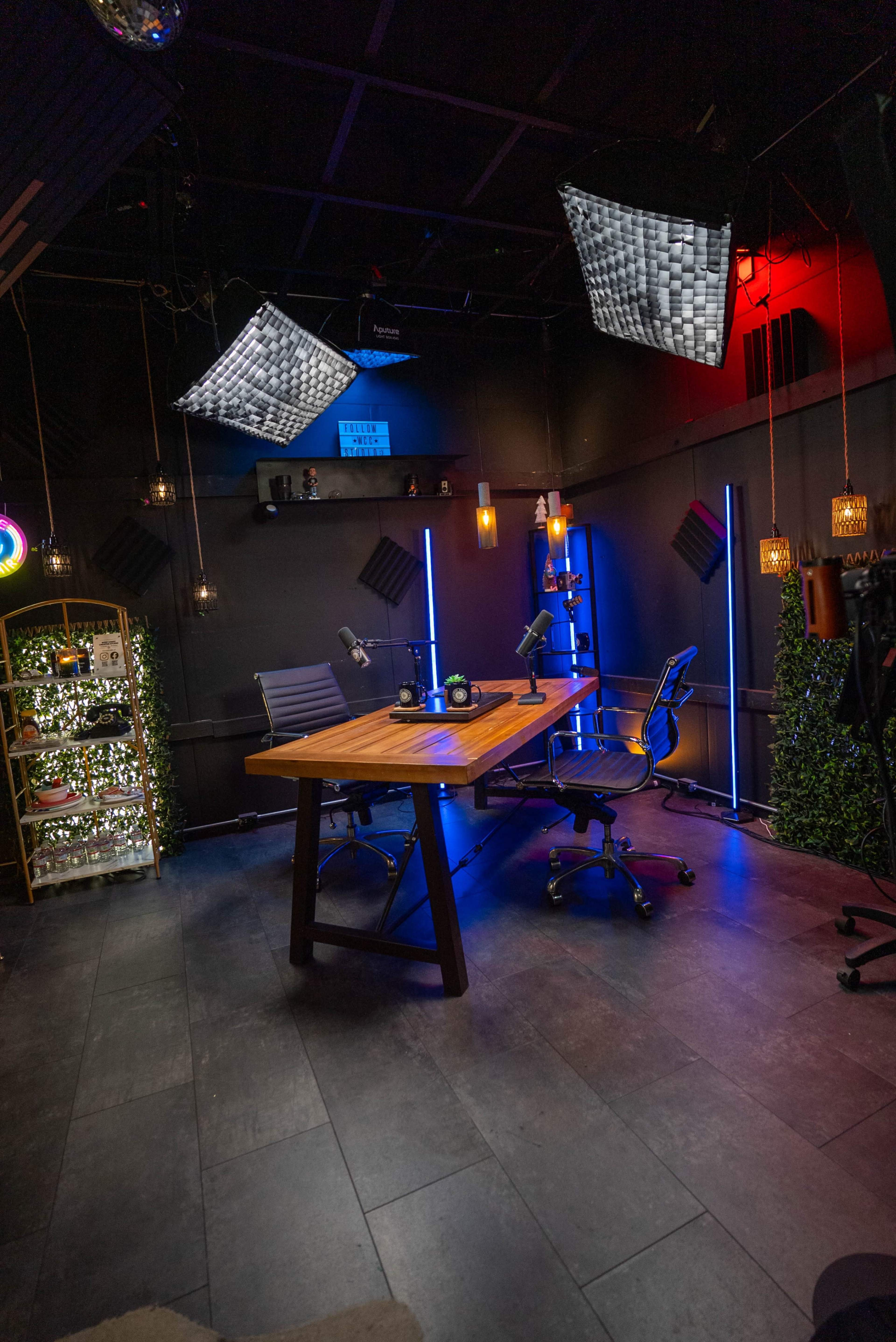 The image shows a modern podcast or video recording studio with a wooden table, two chairs, microphones, and various decorative lighting elements.