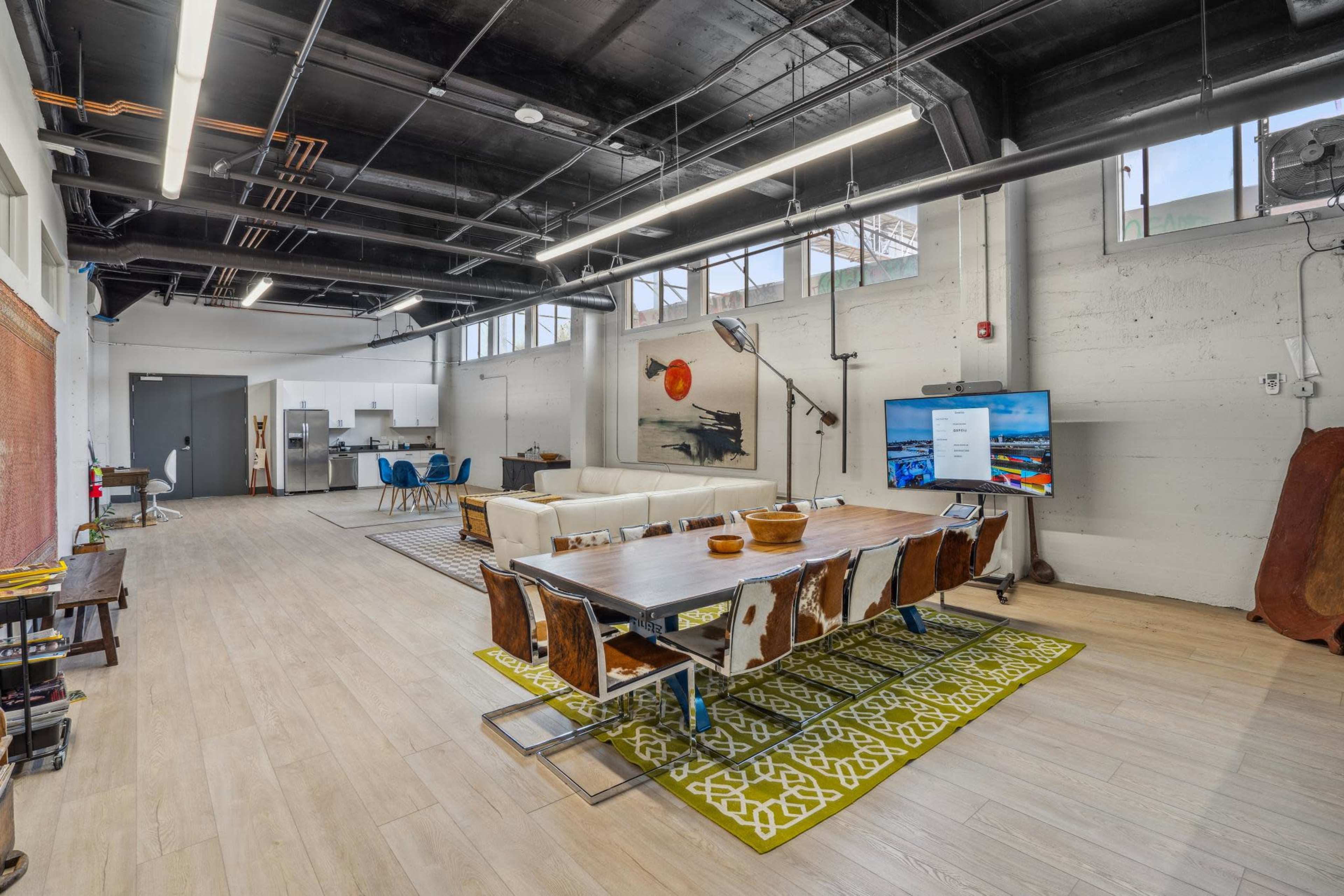 The image shows a modern open-concept living space with a dining area featuring a long table, mixed seating, and a large television, alongside a kitchen in the background.
