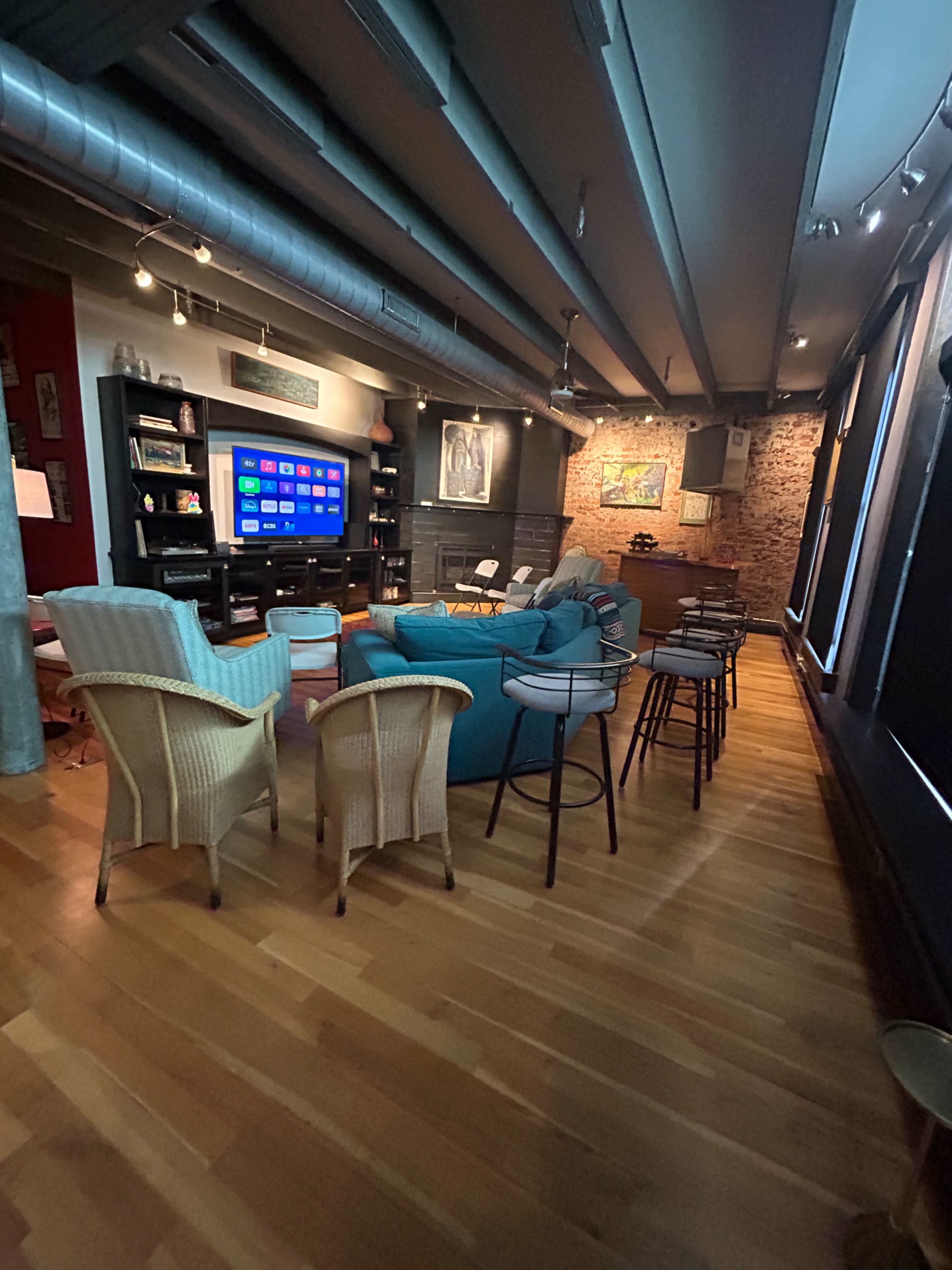 The image depicts a modern entertainment room featuring a turquoise sofa, various seating options including chairs and bar stools, and a large wall-mounted TV surrounded by shelves.