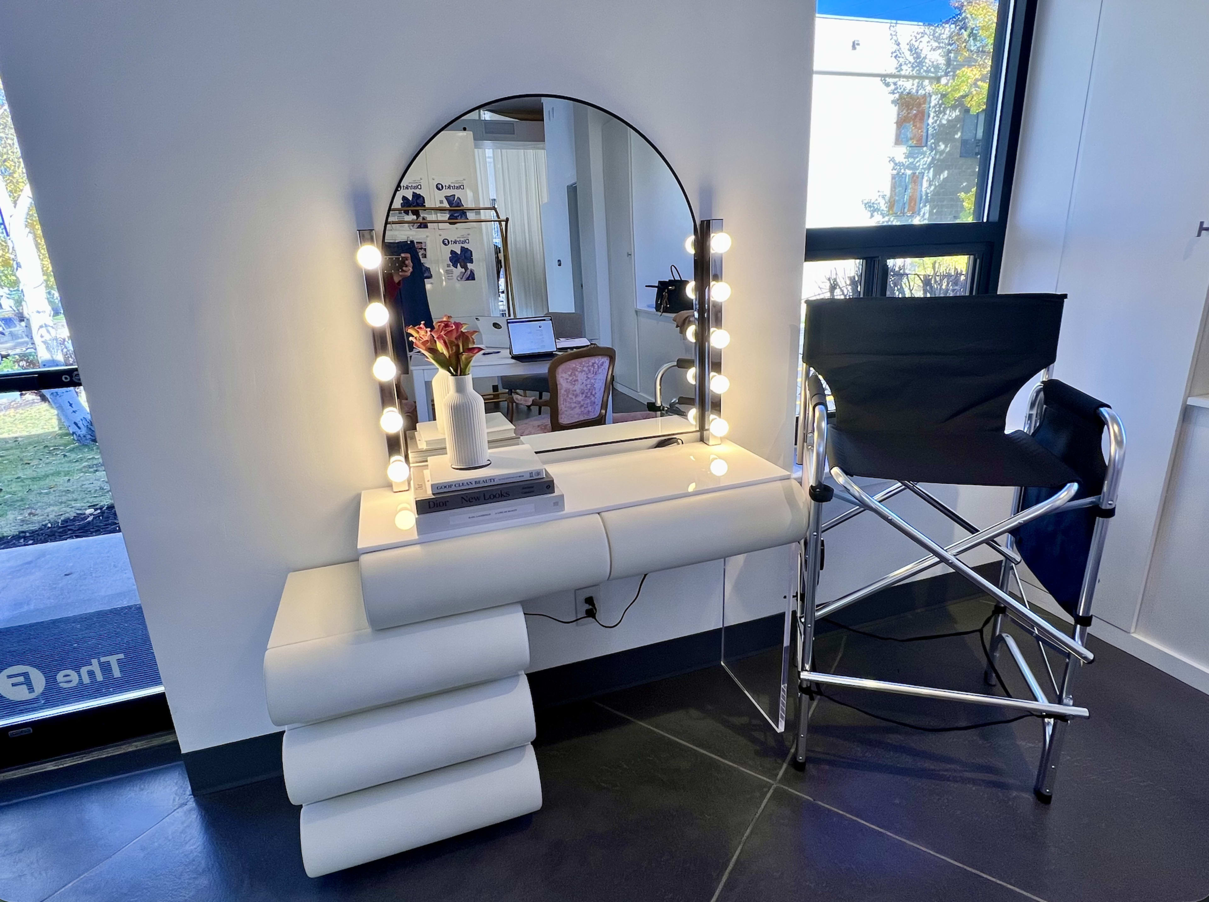 A minimalist vanity setup features a large round mirror with bright bulbs, a sleek white countertop, a stack of decorative books, a small vase with flowers, and a black folding director's chair beside it.
