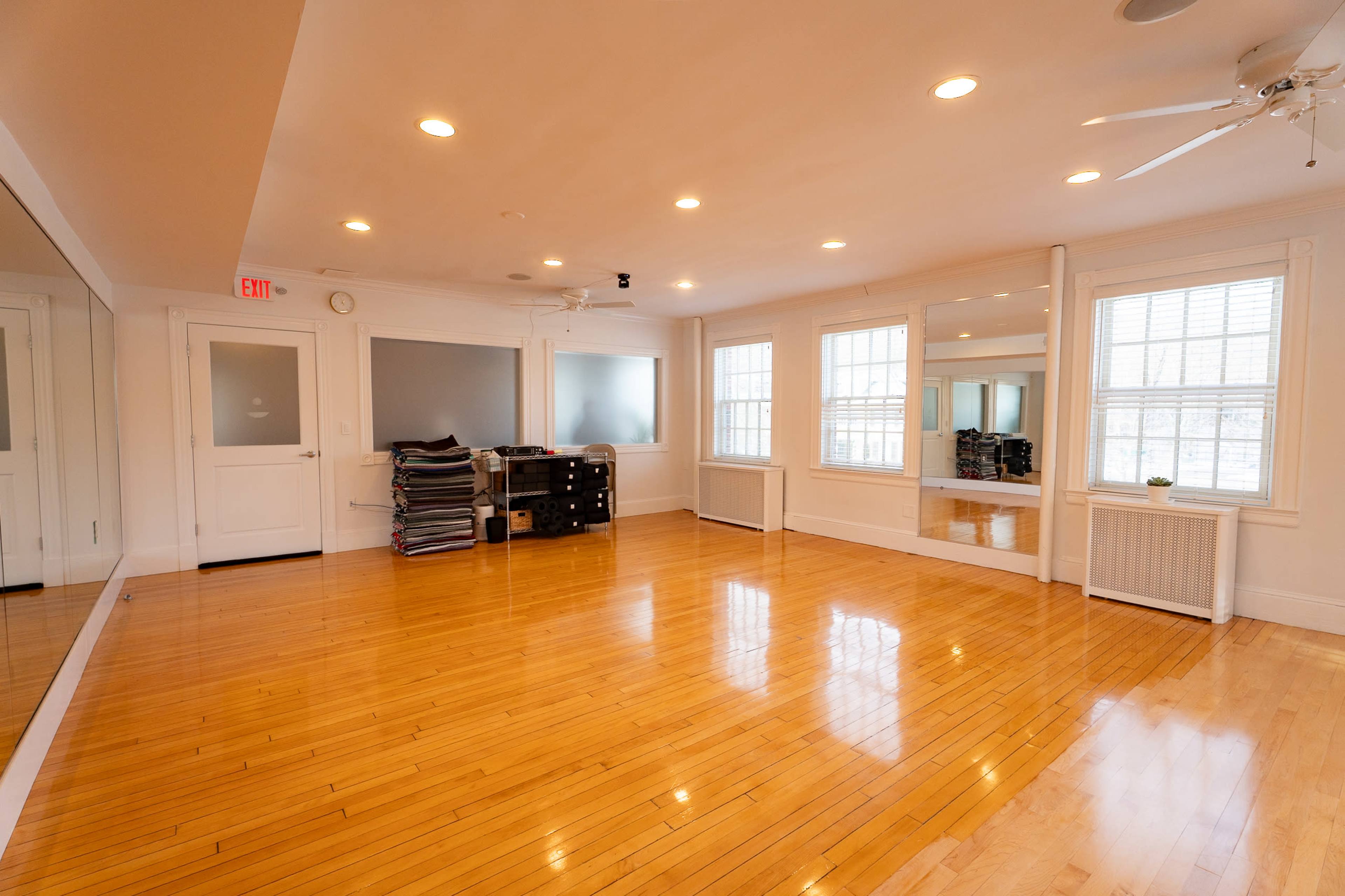 The image shows a spacious, well-lit room with polished wooden floors, large windows, and stacks of folded materials on one side.