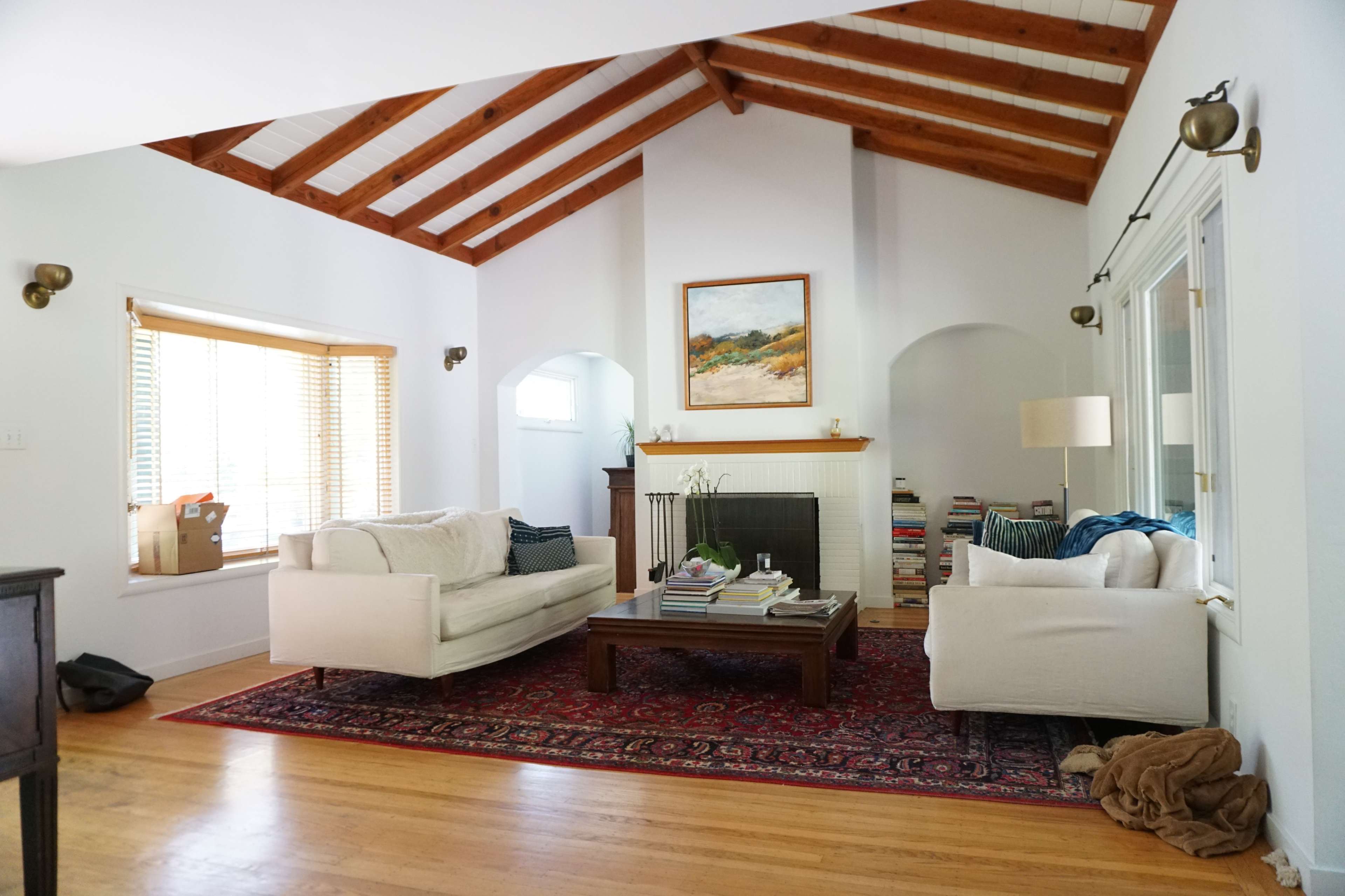 A spacious living room features a vaulted ceiling with wooden beams, two white sofas, a wooden coffee table piled with books, and large windows with brown blinds.