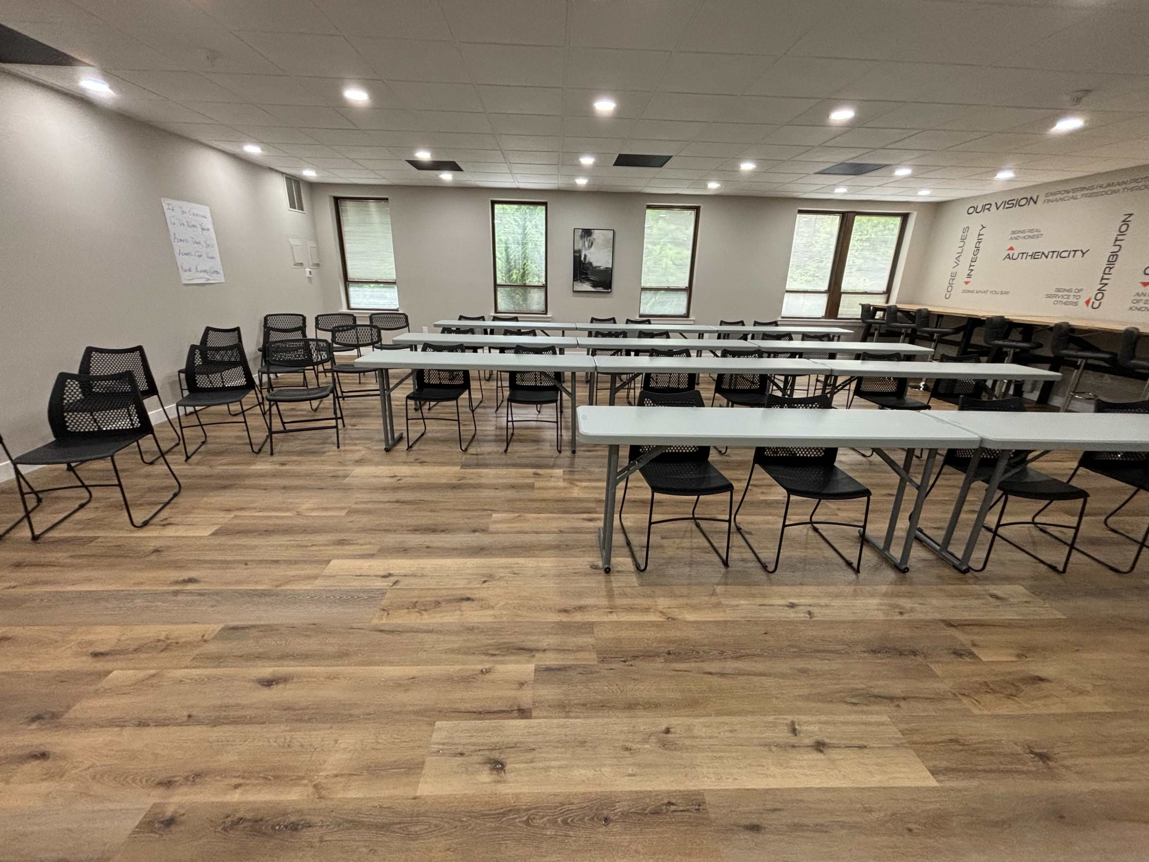 A spacious meeting room arranged with several tables and chairs, featuring large windows that allow natural light to enter.