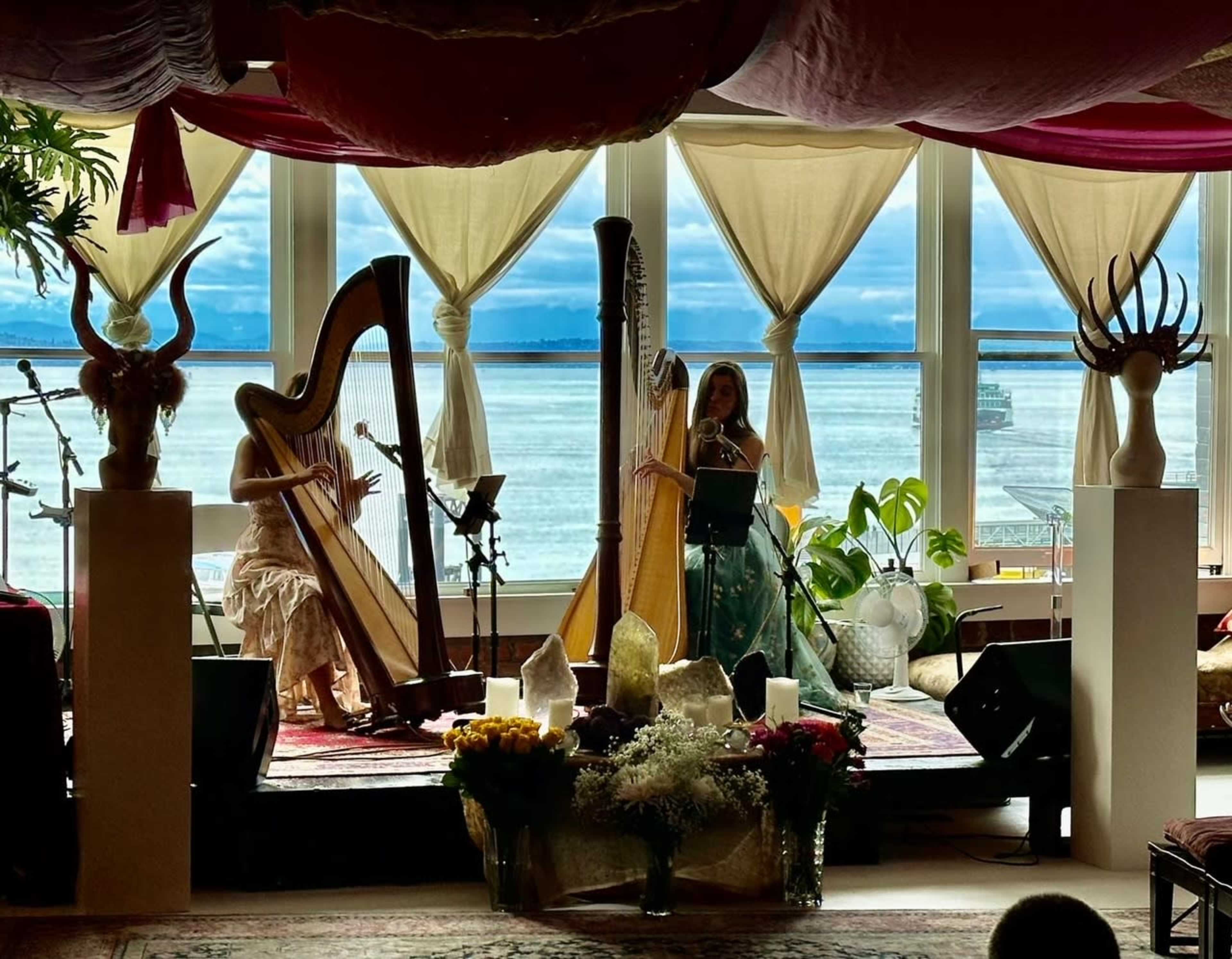 Two musicians play harps on a stage adorned with plants and decorative elements, set against a backdrop of large windows overlooking a body of water.