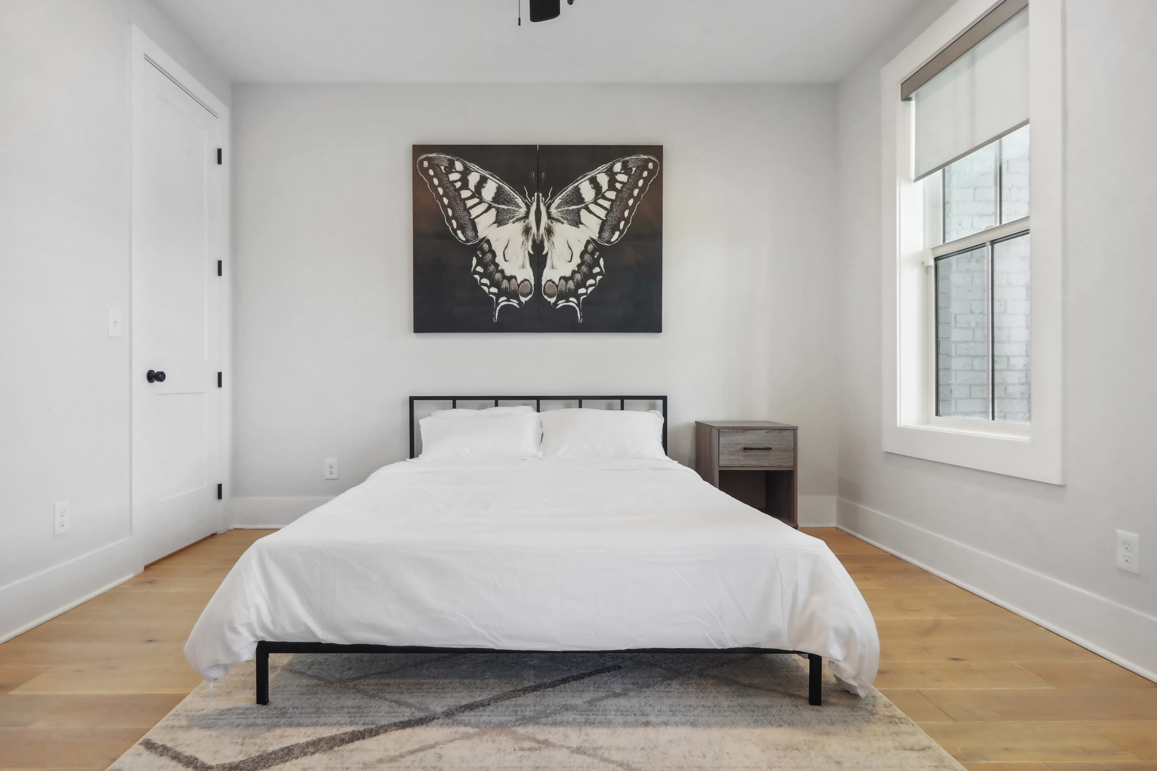 A minimalist bedroom features a bed with a white bedspread, a nightstand, and a large butterfly artwork on the wall.