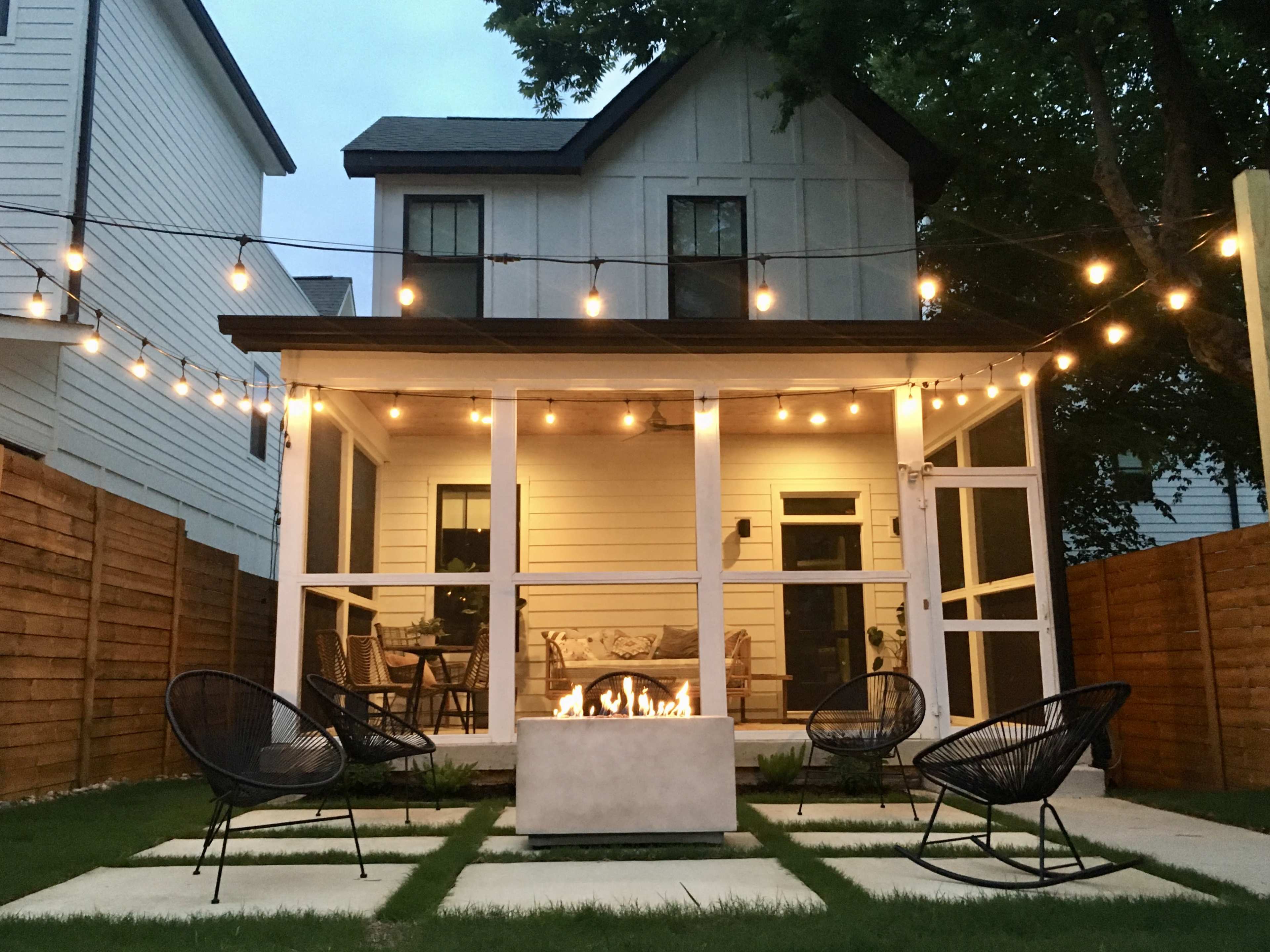 A backyard patio features a fire pit surrounded by four chairs, with string lights overhead illuminating the space and a house in the background.