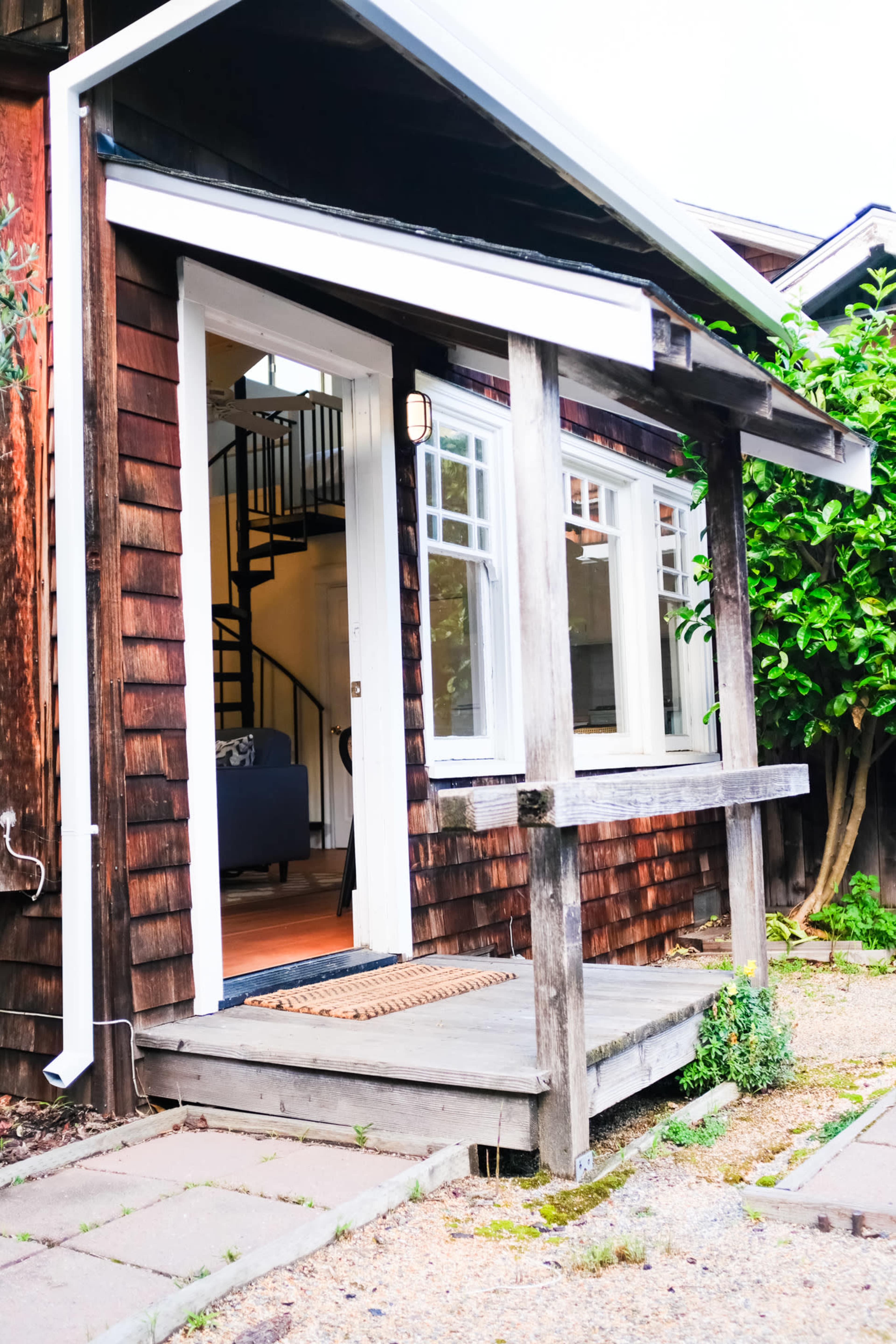 A small wooden house with a front porch, leading to a doorway that opens into an interior space, with a staircase visible through the window.