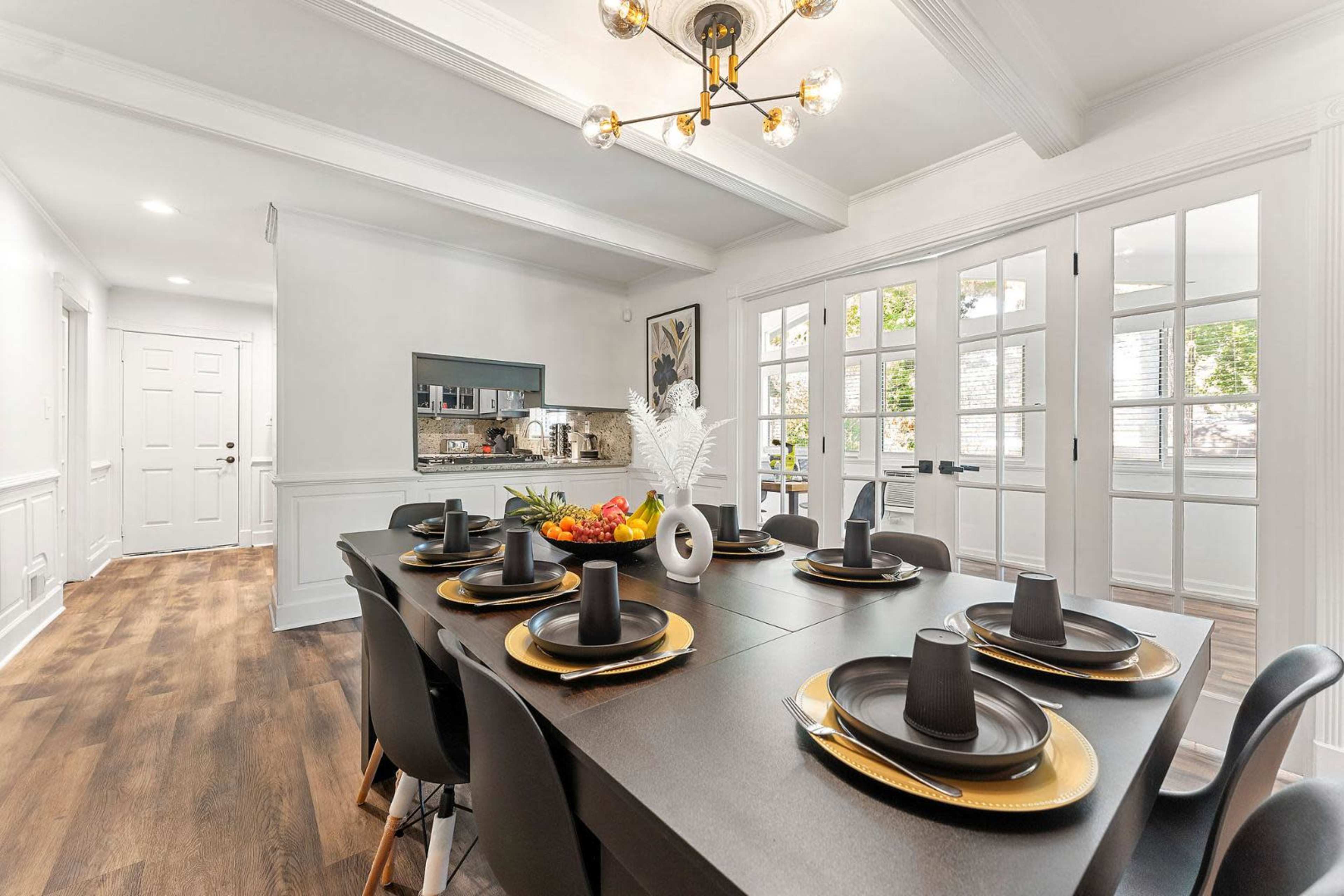 A modern dining area features a long black table set with plates and cups, surrounded by black chairs, with a kitchen visible in the background.