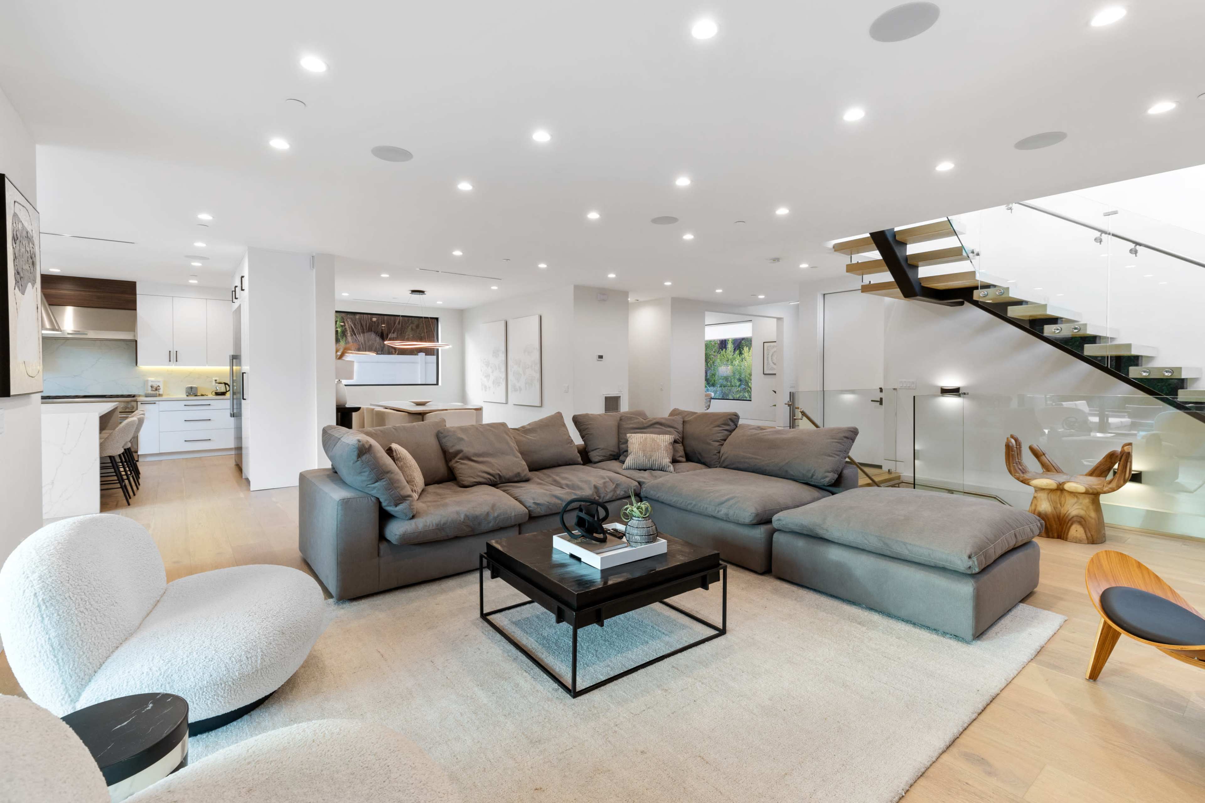 The image shows a modern living room featuring a large sectional sofa, a coffee table, and a staircase with glass railings leading to an upper level.