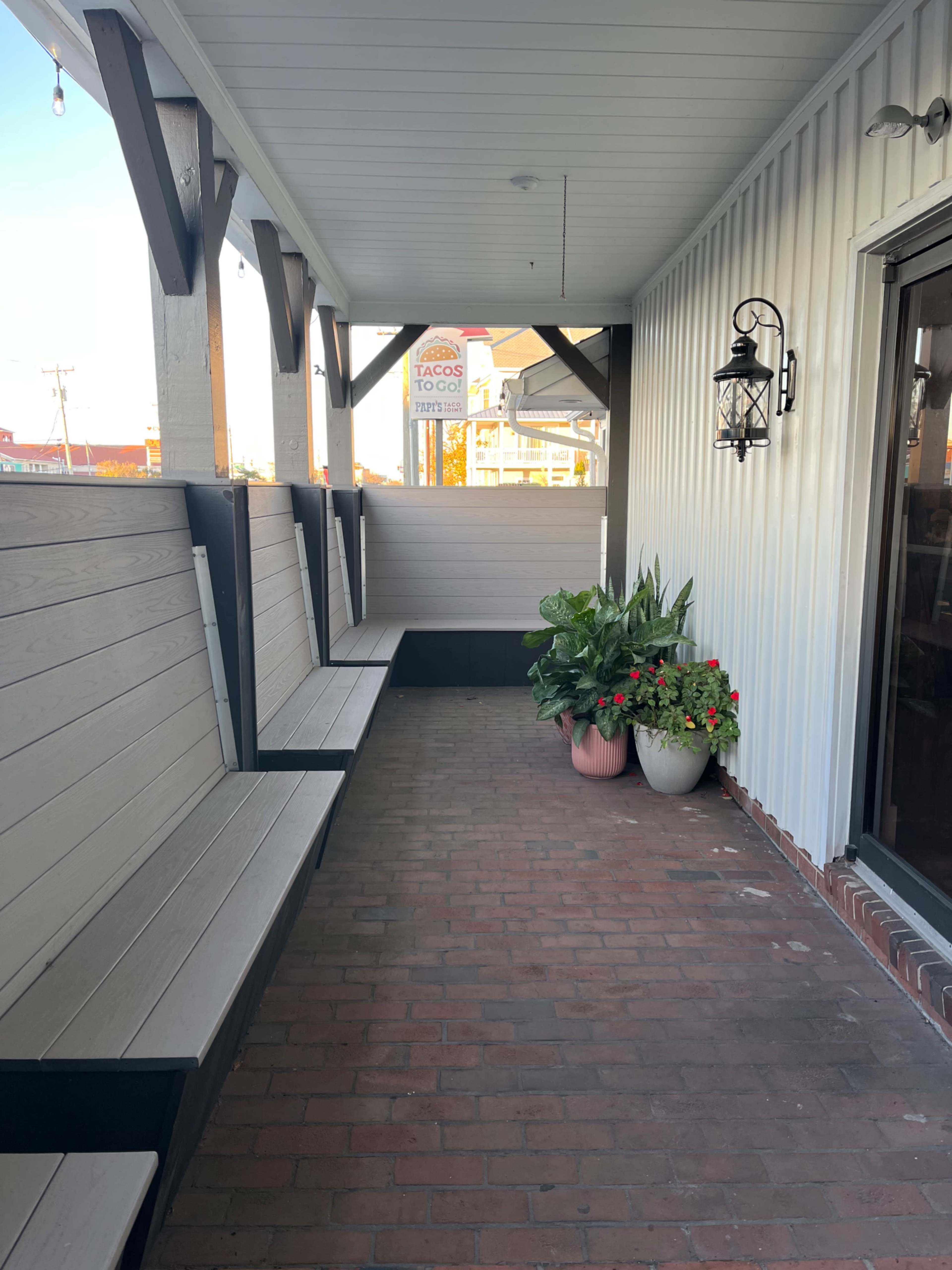 A covered porch area with brick flooring, featuring bank-style seating along the walls and potted plants near the entrance.