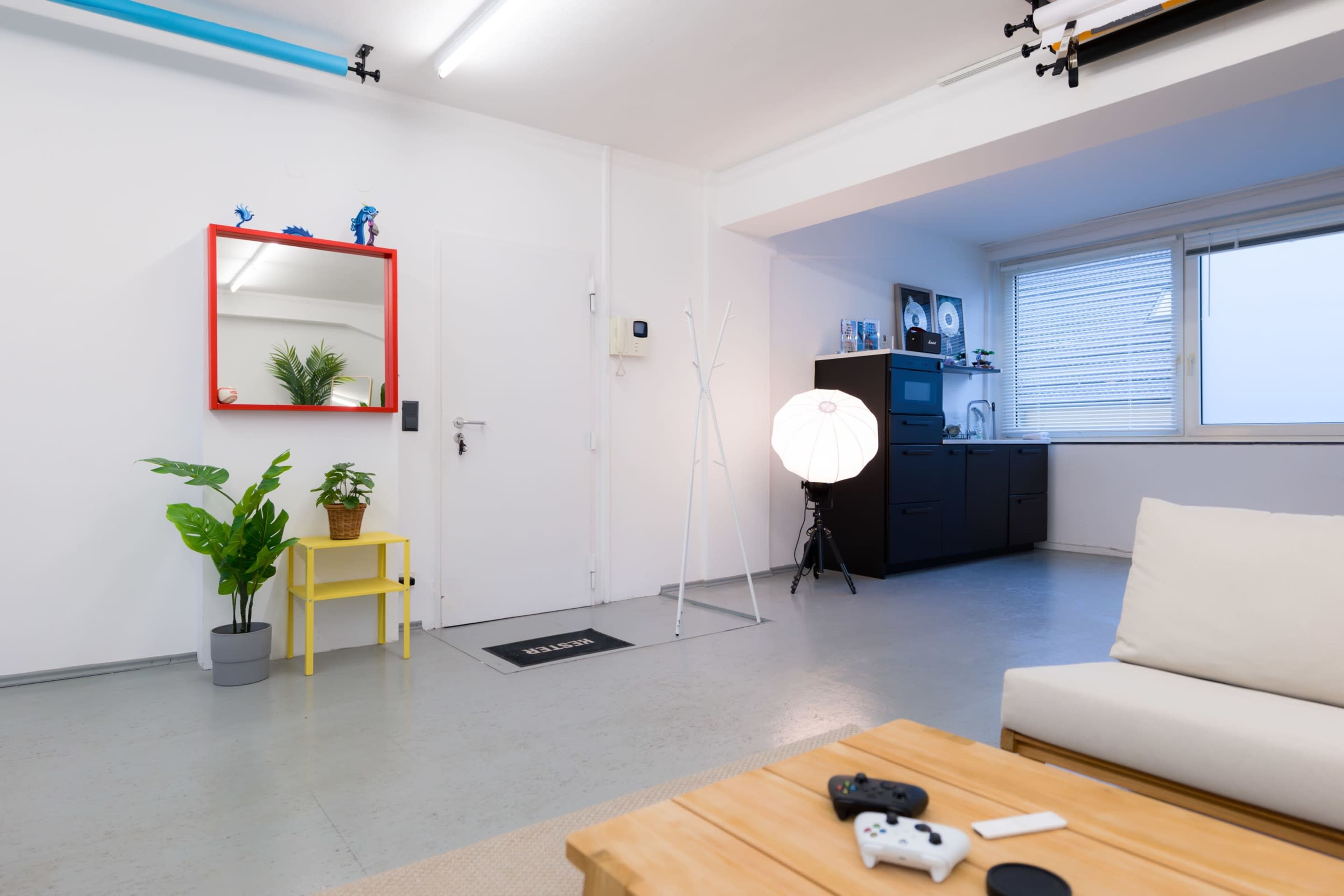 The image shows a modern, minimalistic interior featuring a living area with a white sofa, a wooden coffee table, a plant in a yellow pot, and a door leading to another room.