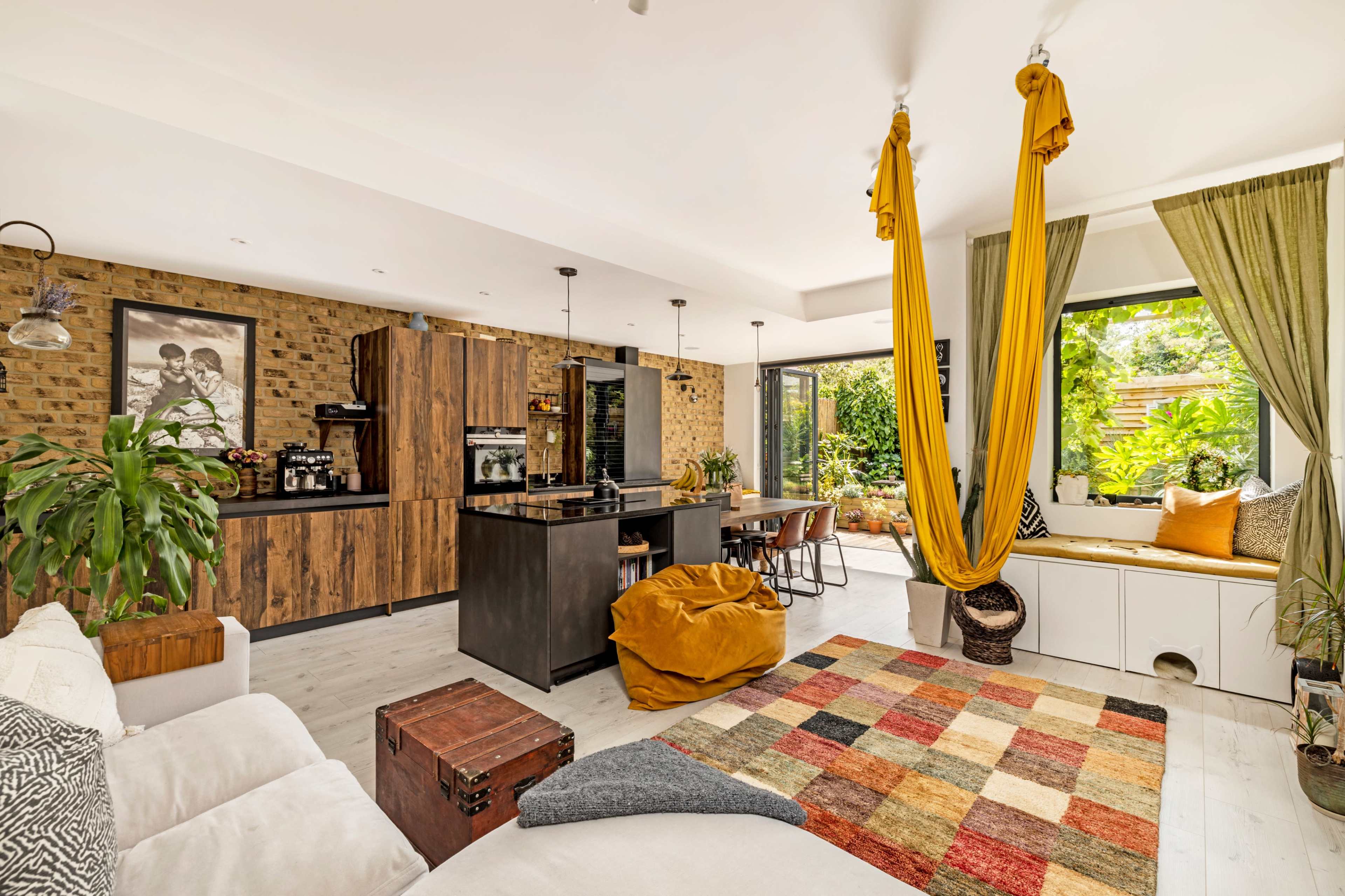 A modern living space featuring a kitchen, dining area, and a cozy seating area, with large windows that reveal a green outdoor garden.