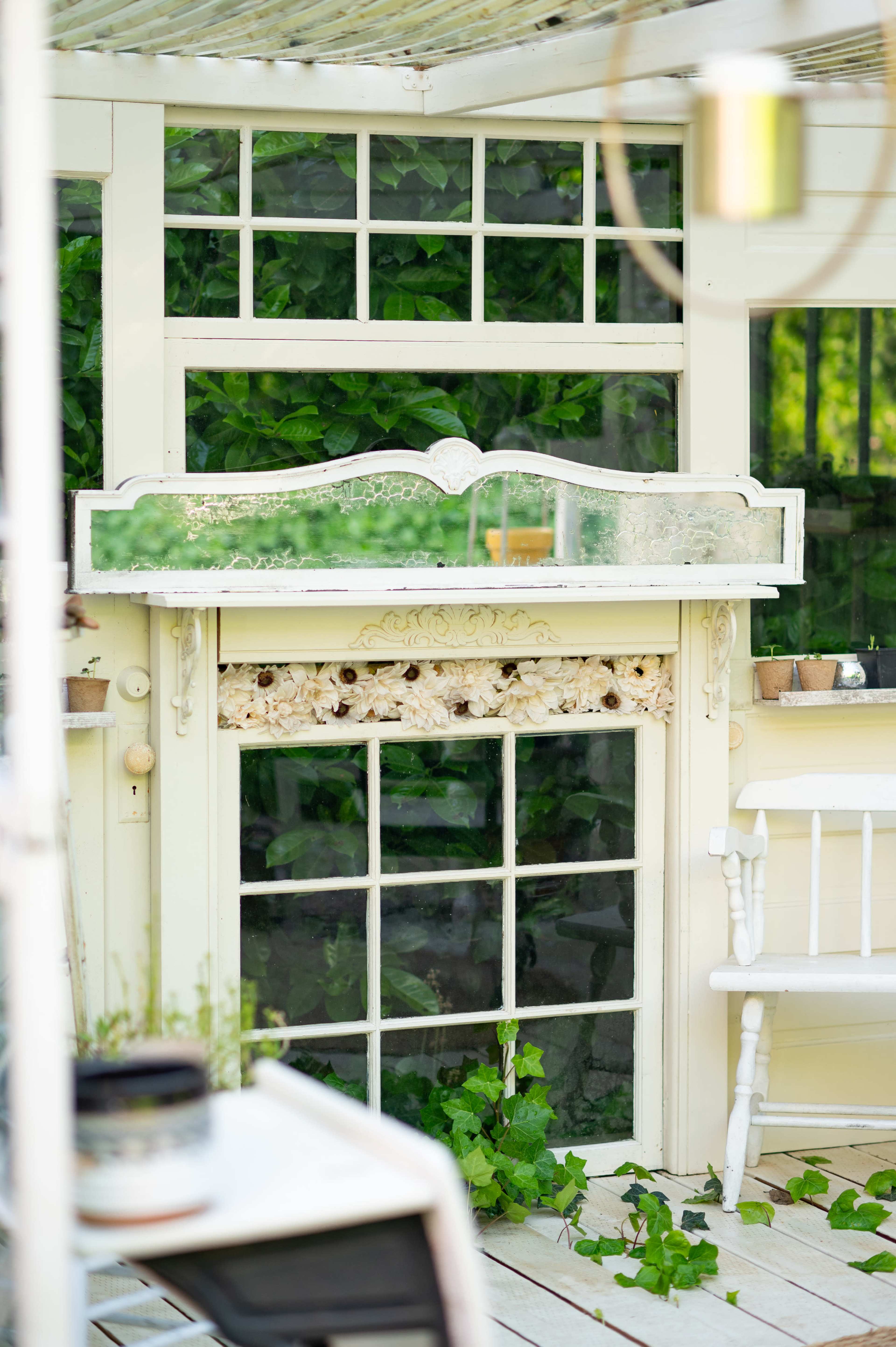 The image shows a bright indoor space featuring a vintage wooden mantelpiece against a backdrop of greenery visible through large windows.