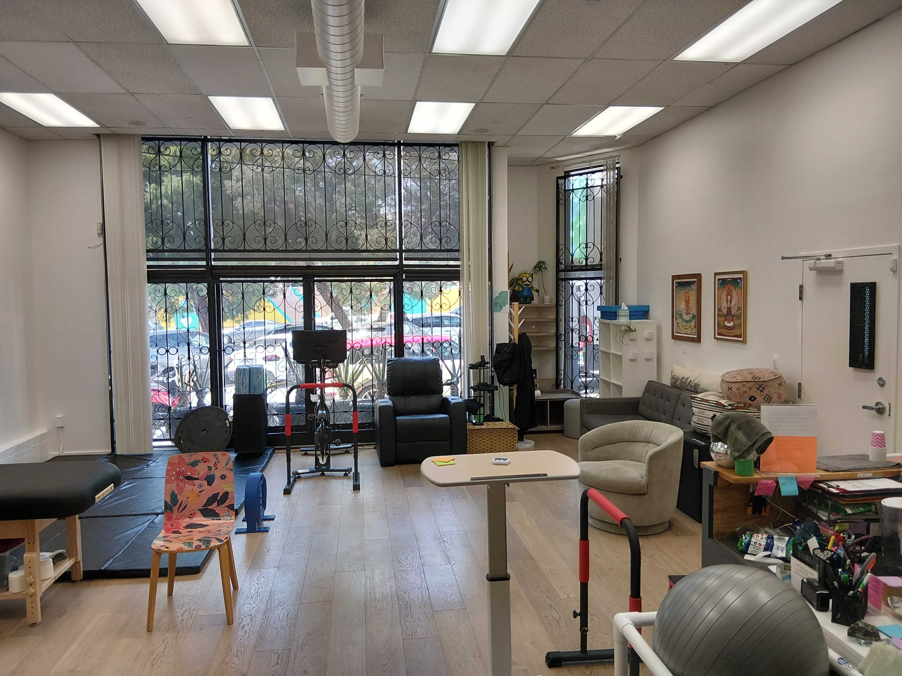 The image shows a spacious room designed for therapy or rehabilitation, furnished with various seating options, exercise equipment, and large windows.
