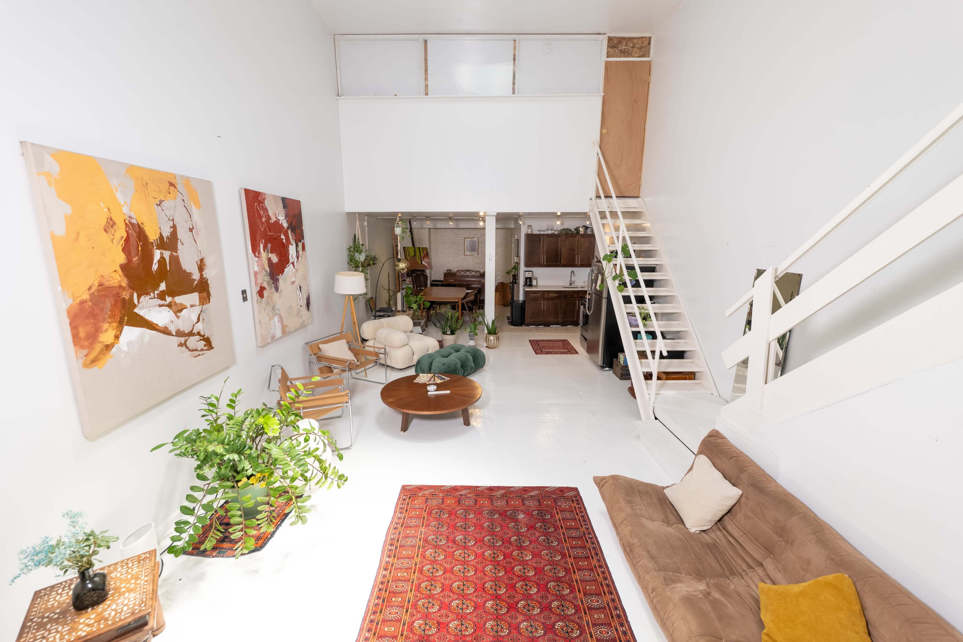 A spacious, minimalist living area featuring a brown sectional sofa, a round coffee table, vibrant artworks on the walls, and a staircase leading to an upper level.
