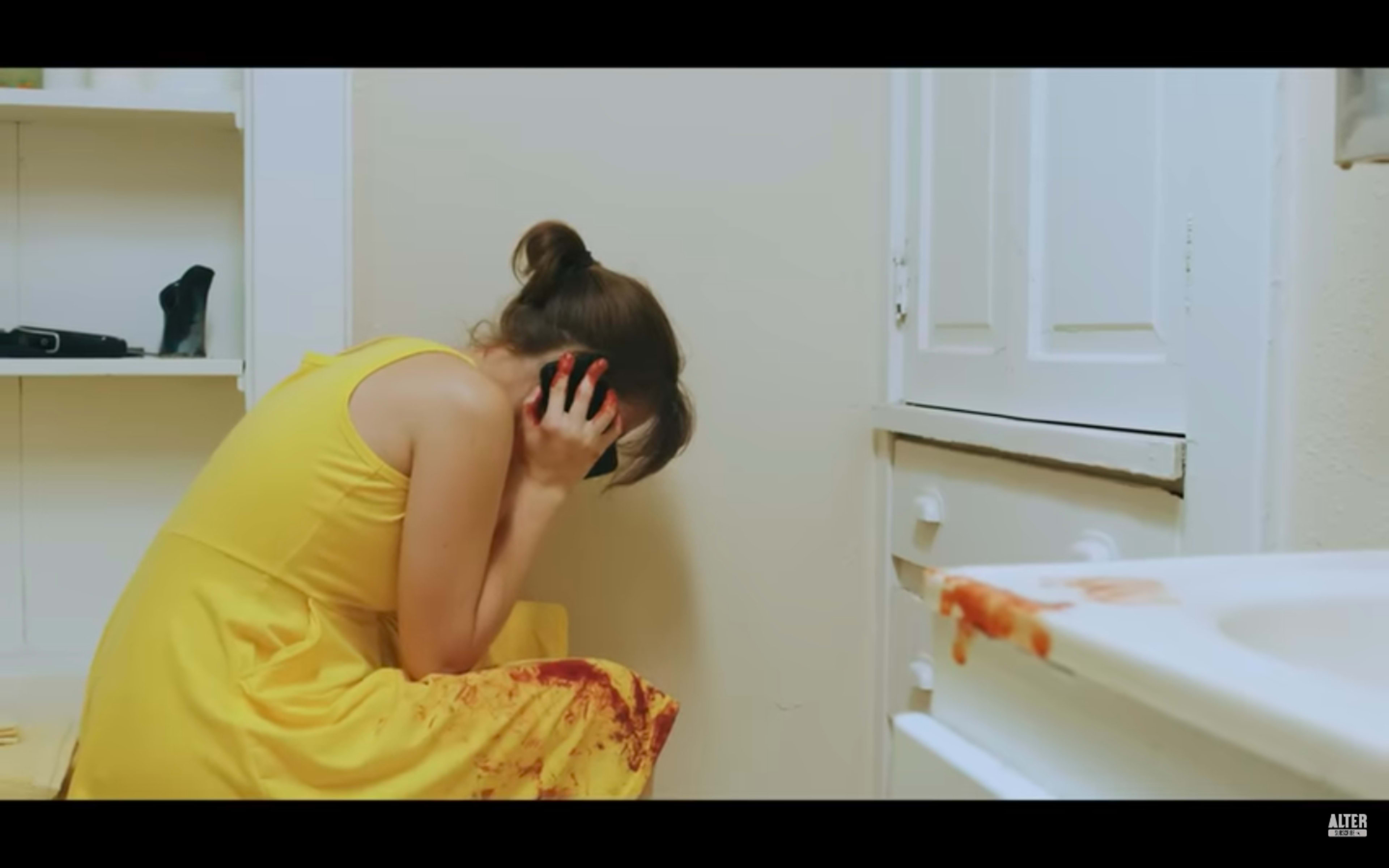 A woman in a yellow dress sits on the floor of a bathroom, holding her head and appearing distressed, while a trail of red liquid is visible on the sink and the wall nearby.