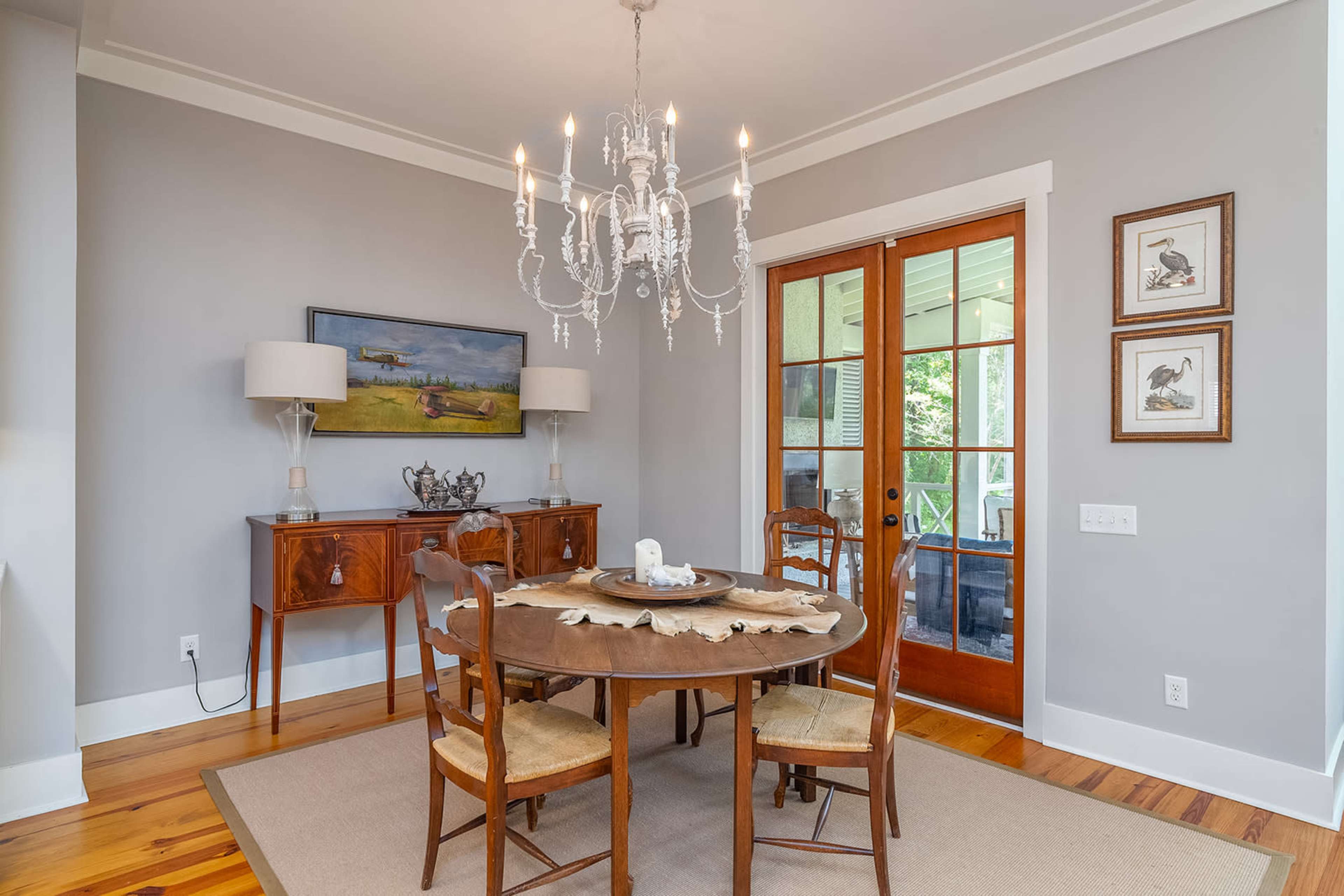 A round dining table with chairs is set under a chandelier in a light-colored room with a sideboard and a door leading to an outdoor area.