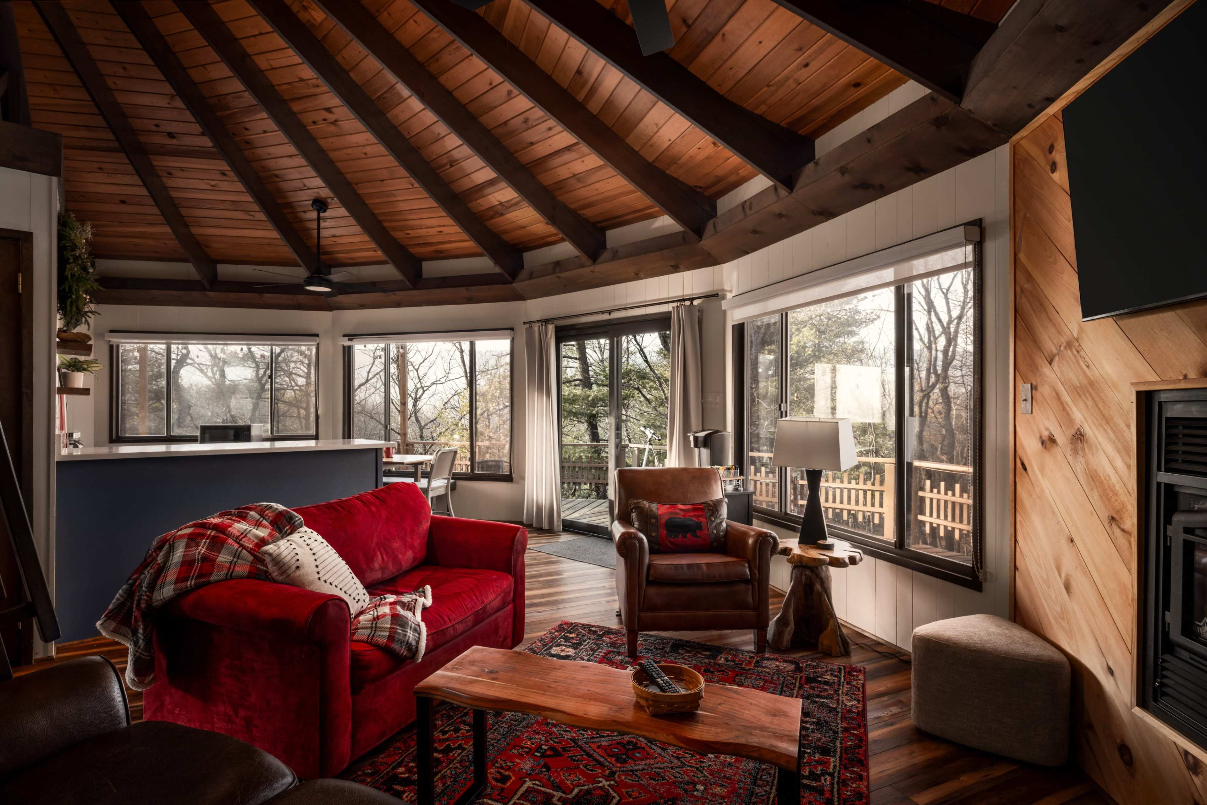 The image shows a cozy living room with a vaulted wooden ceiling, large windows, a red sofa, and a wooden coffee table.