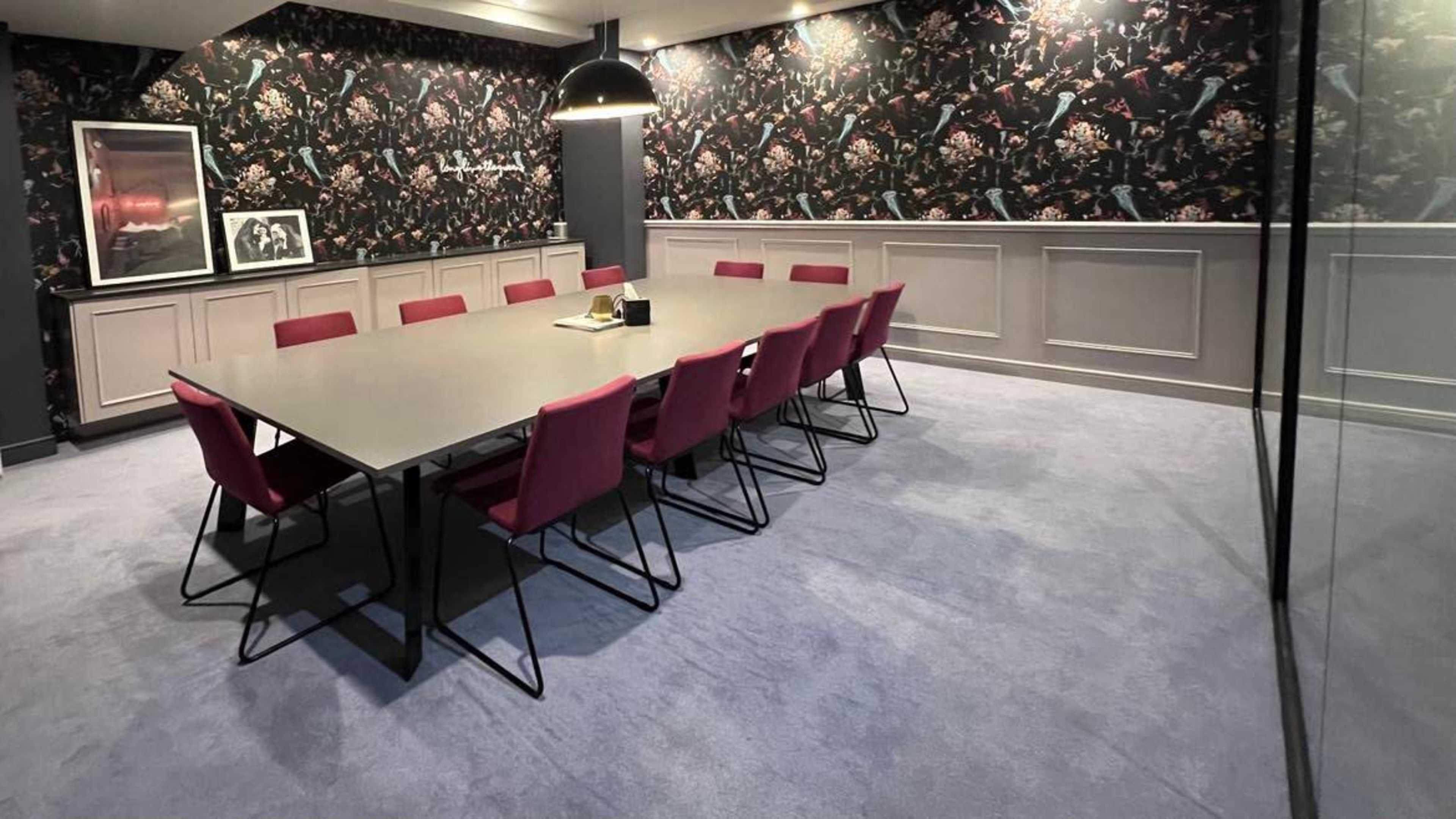 A long conference table surrounded by pink chairs is situated in a modern room with patterned wallpaper and soft carpeting.