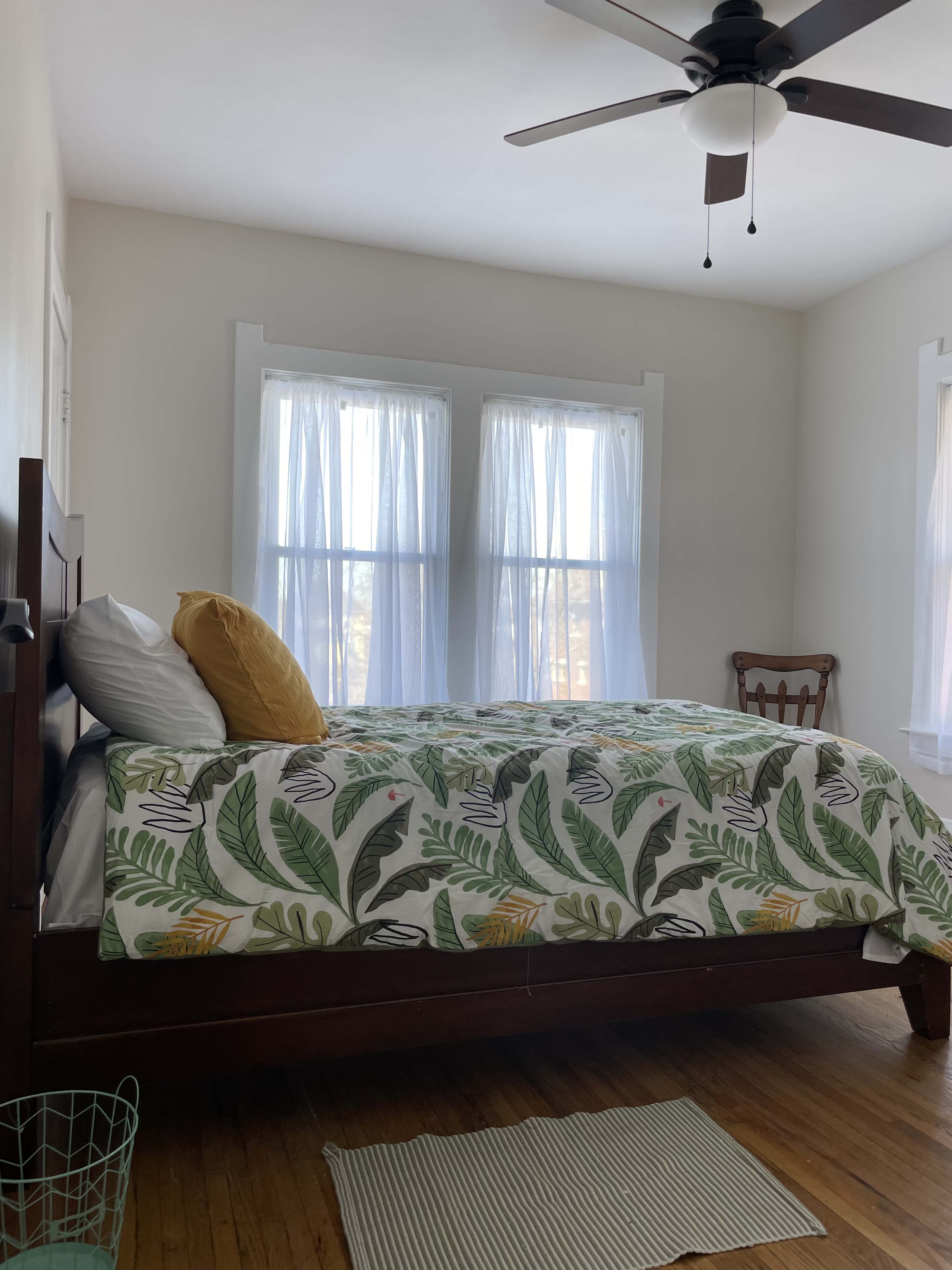 A neatly arranged bedroom with a wooden bed covered in a leafy-patterned quilt, two windows with sheer curtains, and a wooden chair in the corner.