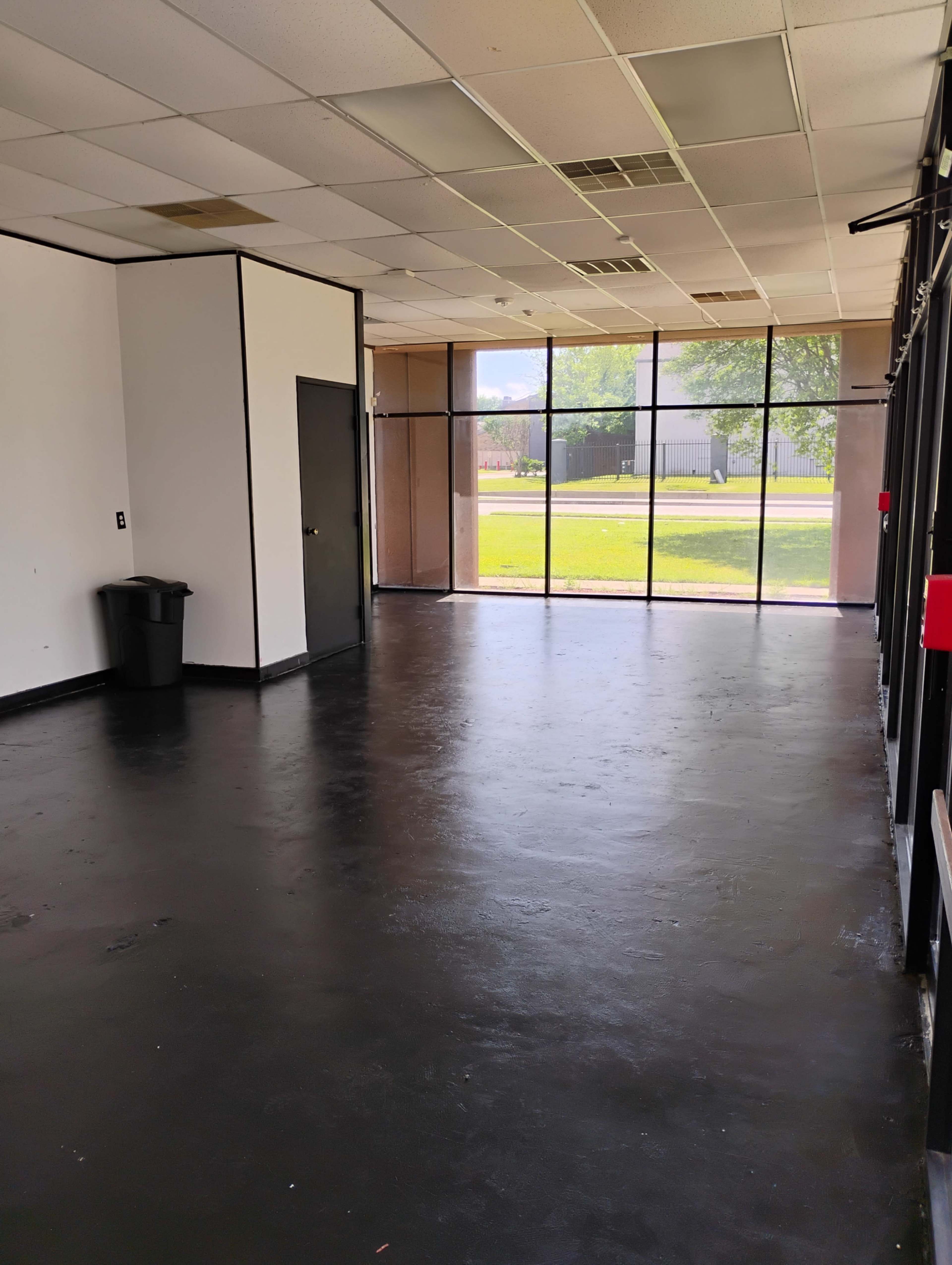 The image shows an empty room with black flooring, a single door, and large windows that provide natural light.